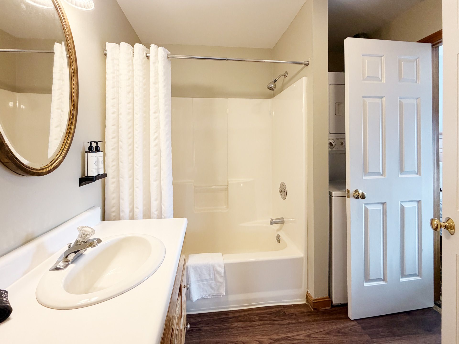 Bright bathroom with white vanity, mirror, bathtub shower, and an open door to a hallway.