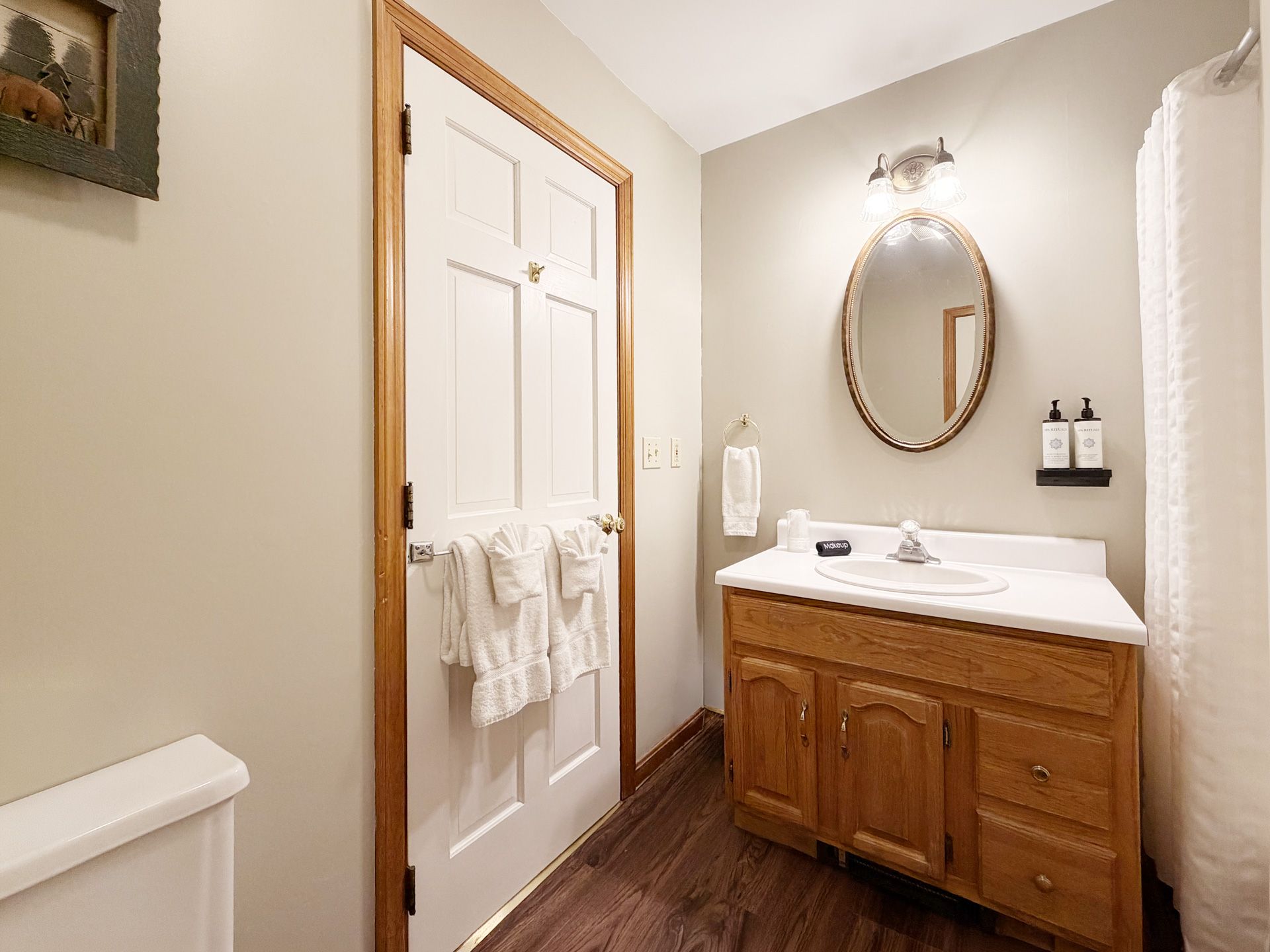 Small bathroom with a wooden vanity, oval mirror, white door, and towel rack.