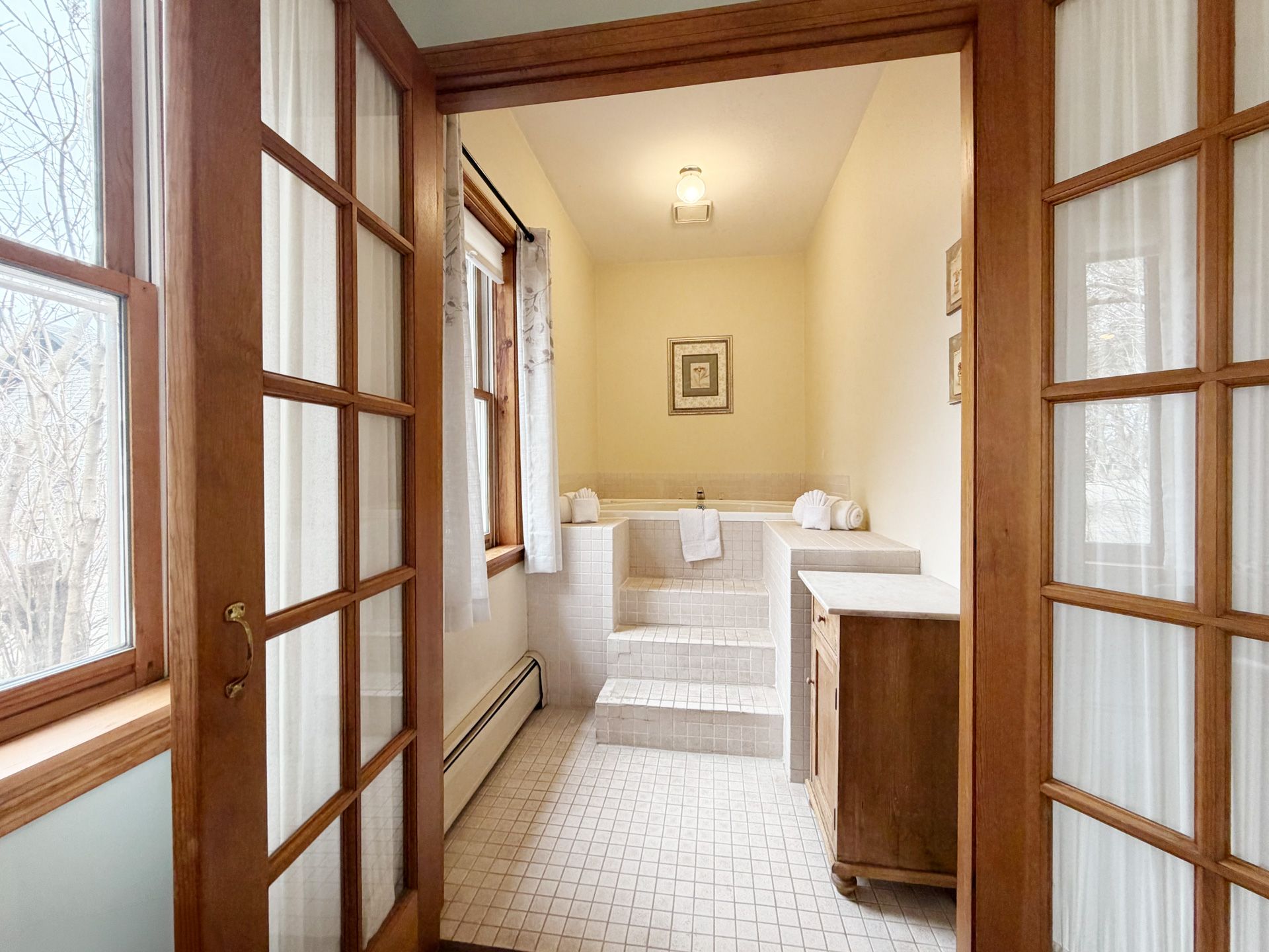 Bright bathroom with white tile, a raised bathtub, wooden cabinets, and French doors leading in.