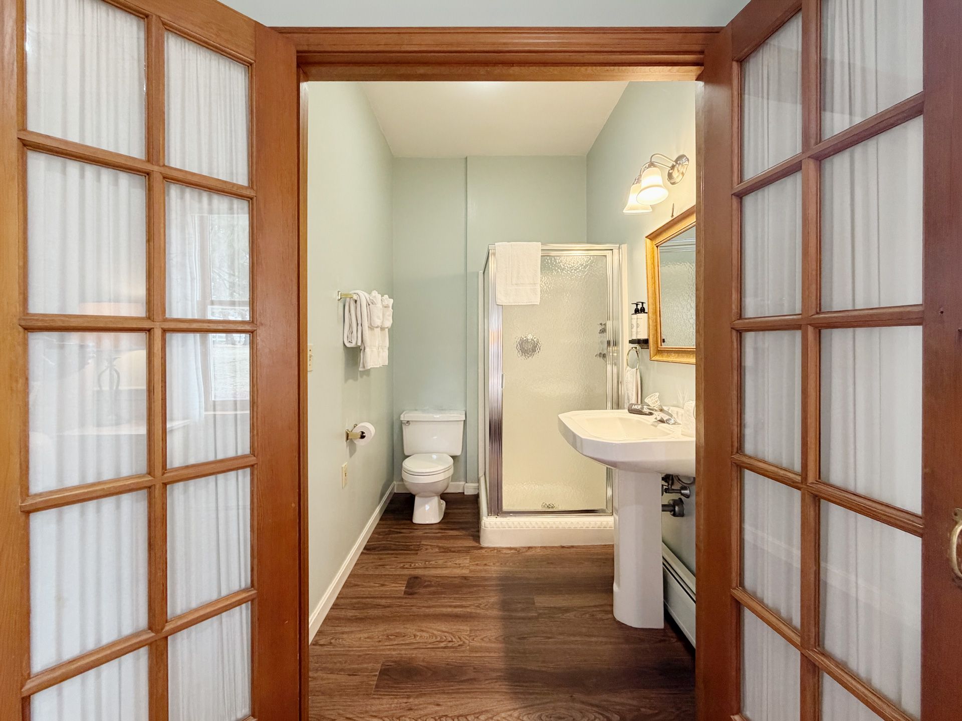 Open wooden doors reveal a small bathroom with toilet, shower, sink, and wood-look floor.