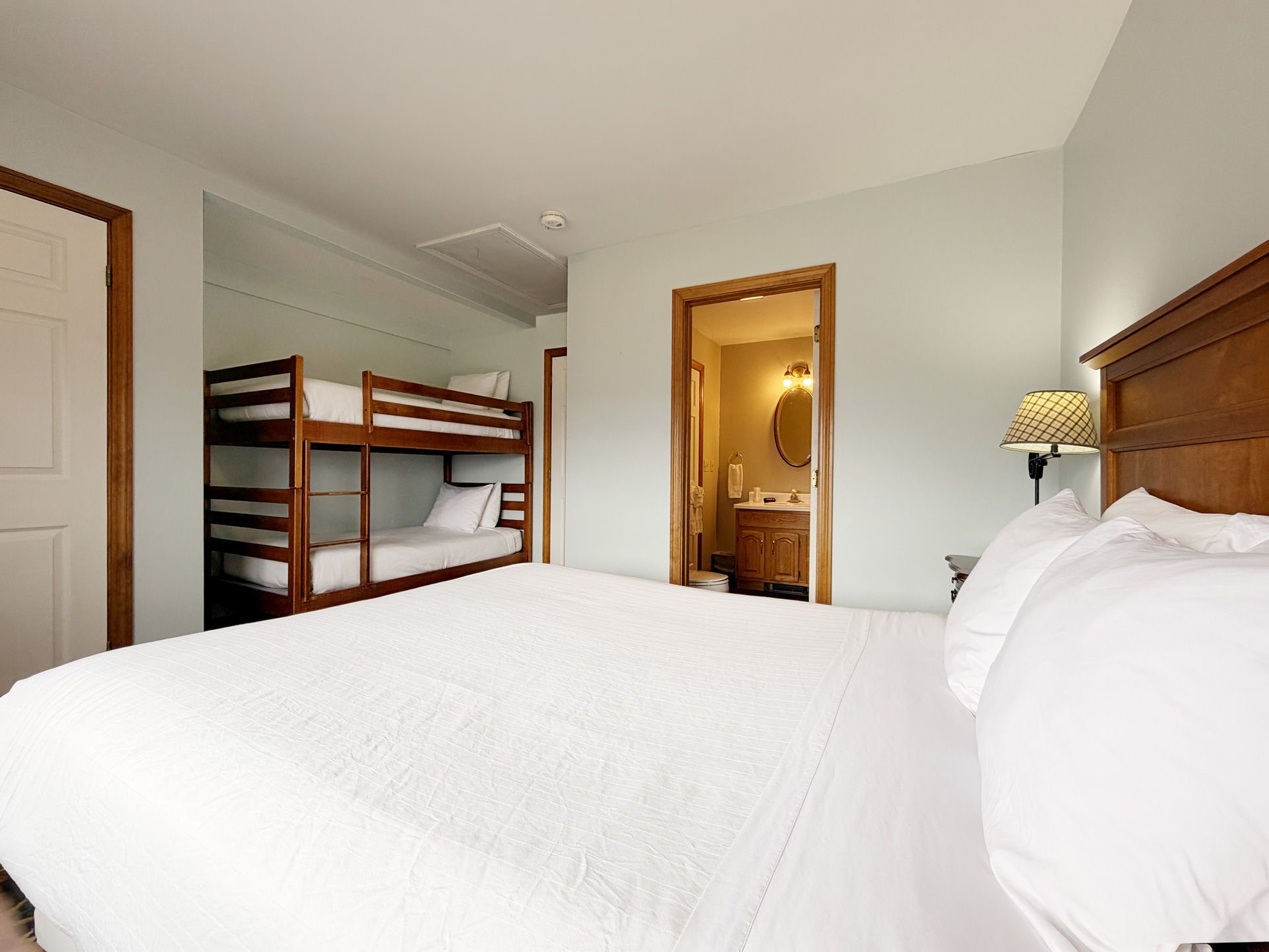 Bright bedroom with white bed, bunk bed, wood door, and dresser lamp