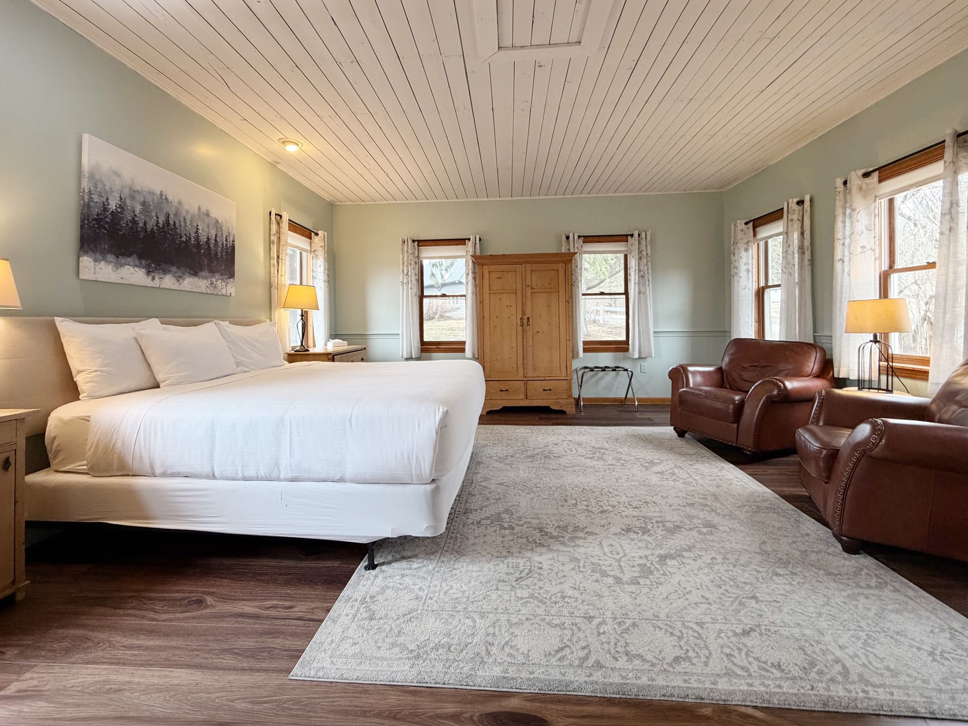 Spacious bedroom with a large white bed, armchairs, dresser, and light gray rug near bright windows.