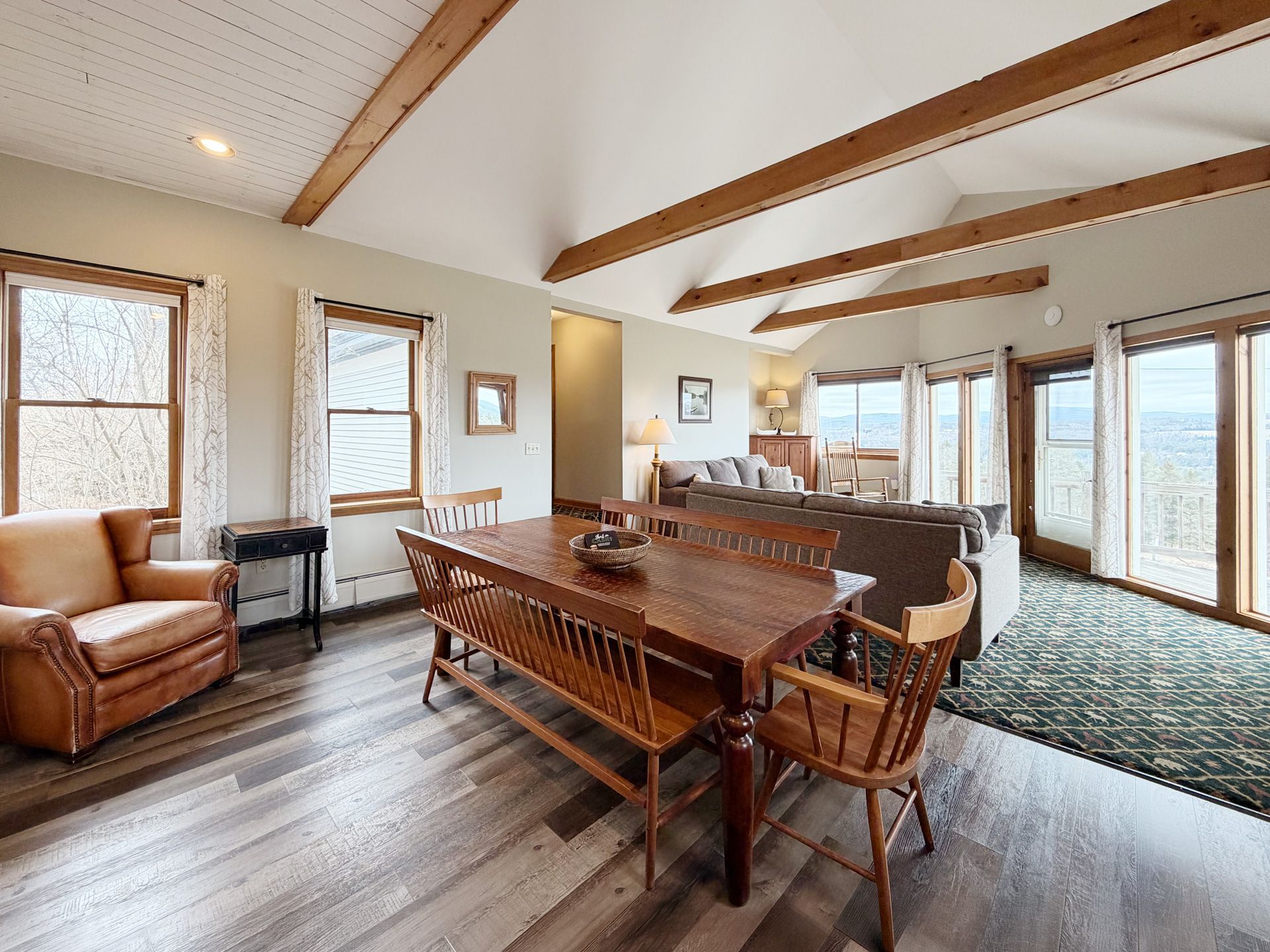 Spacious living room with exposed beams, wood floors, leather chairs, a dining table, and large windows