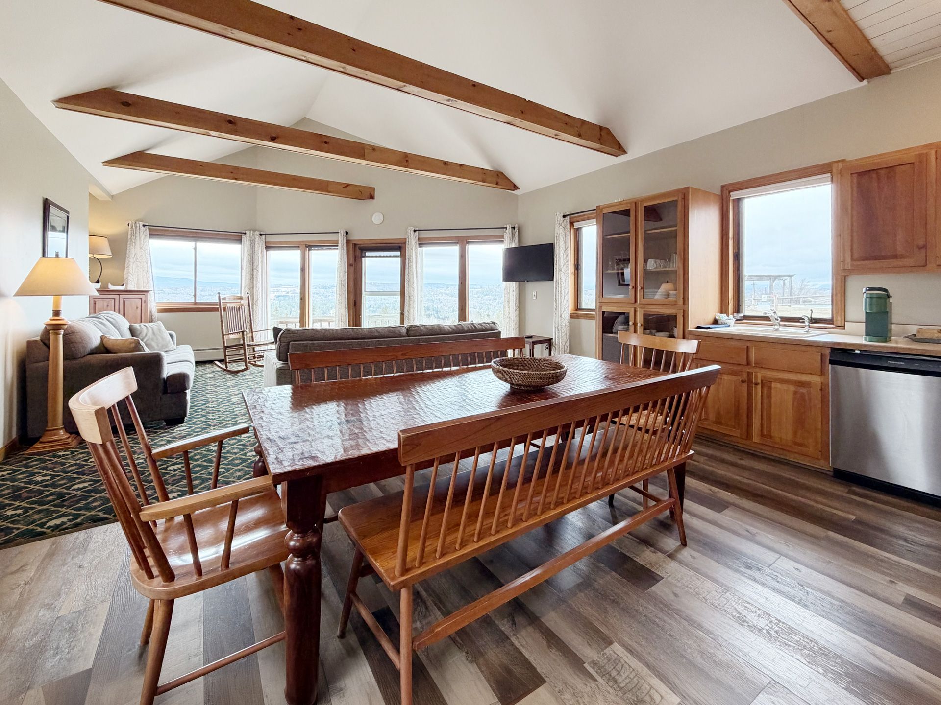 Bright living-dining room with wood beams, dining table, kitchen cabinets, and large windows