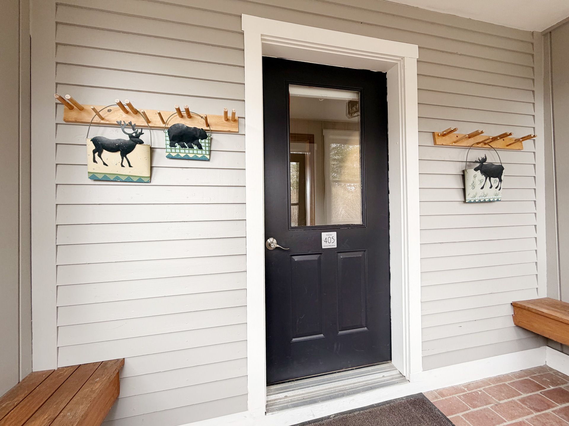 Front porch with a black door, white siding, wooden benches, and small moose wall art.