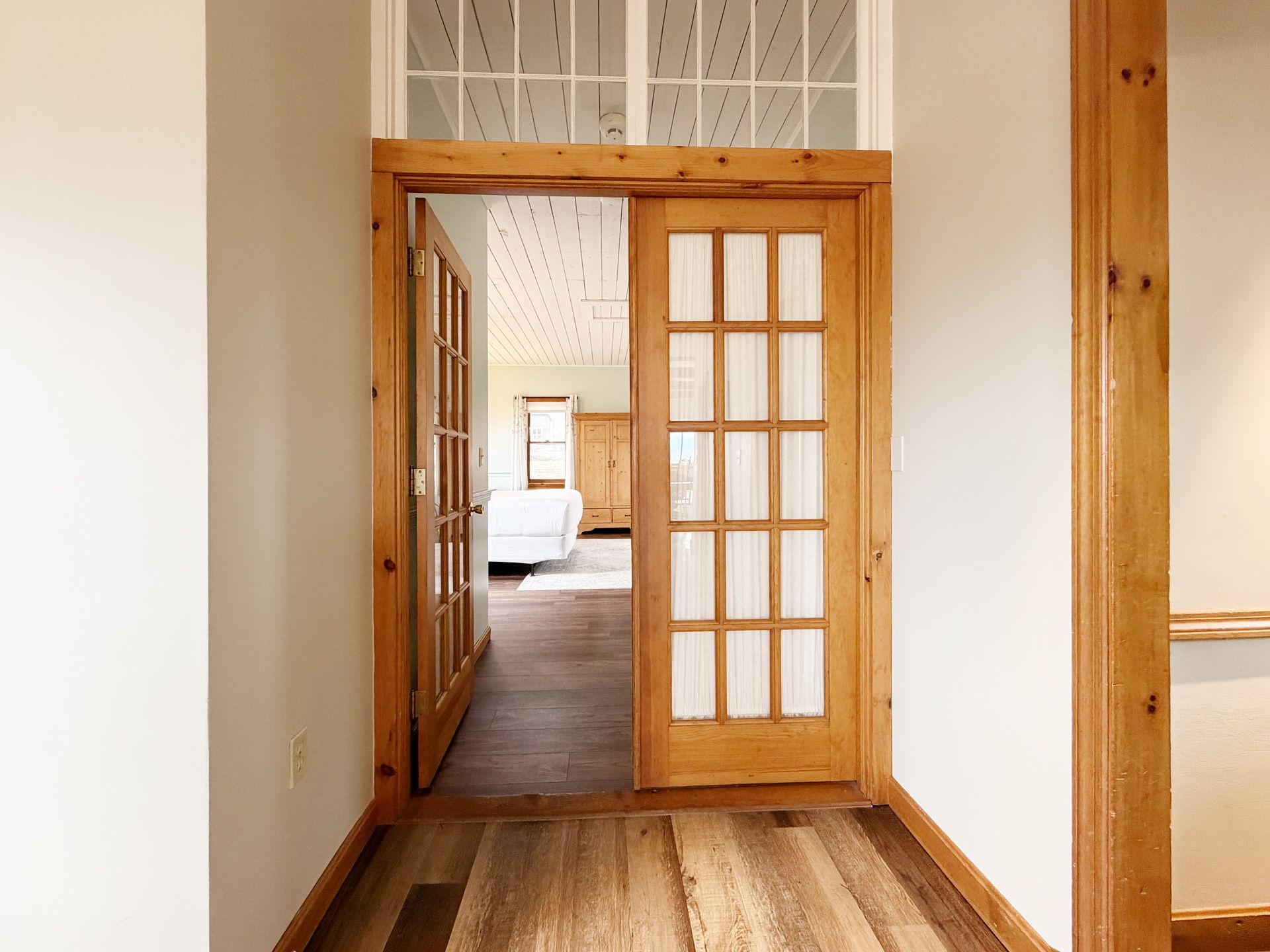 Wood-floored hallway with white walls and a wooden doorway leading to a bright room beyond.