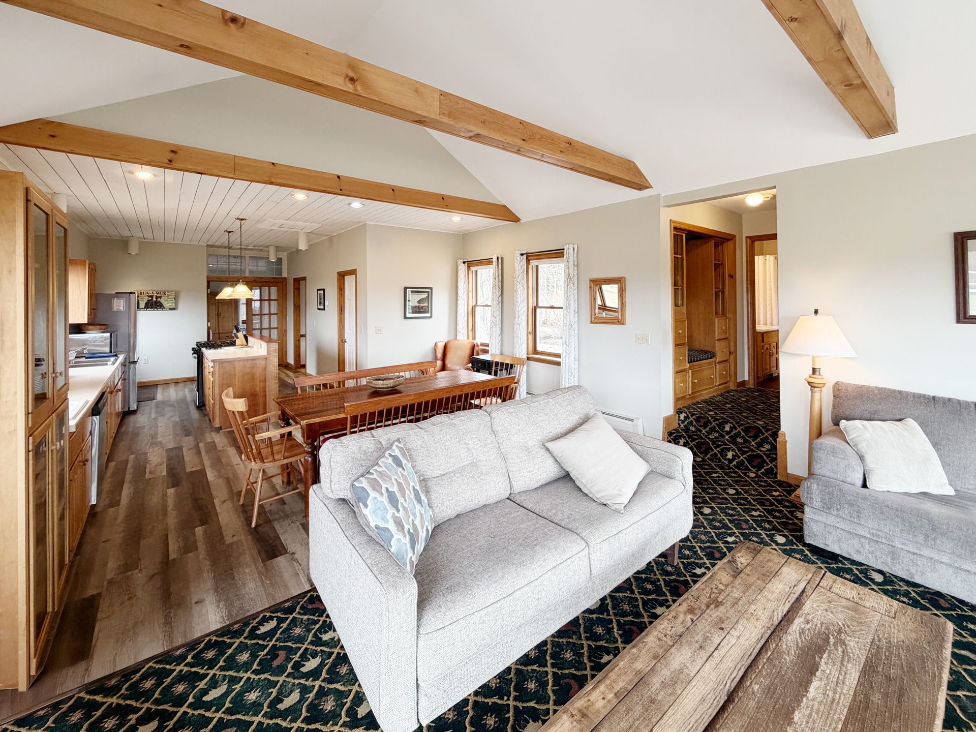 Bright living room with gray sofa, wood beams, patterned rug, and adjacent dining area
