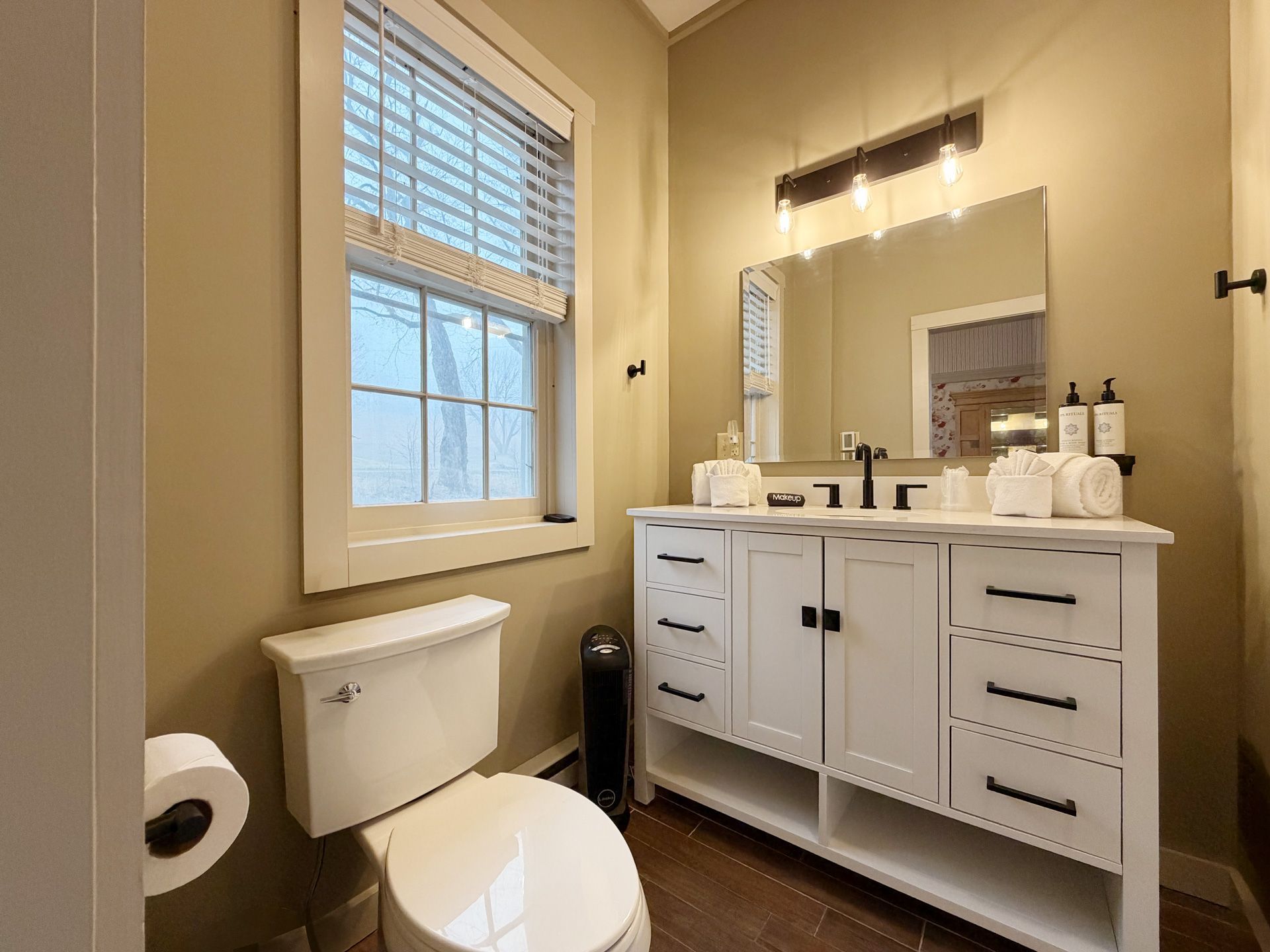 Small beige bathroom with window, white vanity, mirror, sink, toilet, and wall sconces.