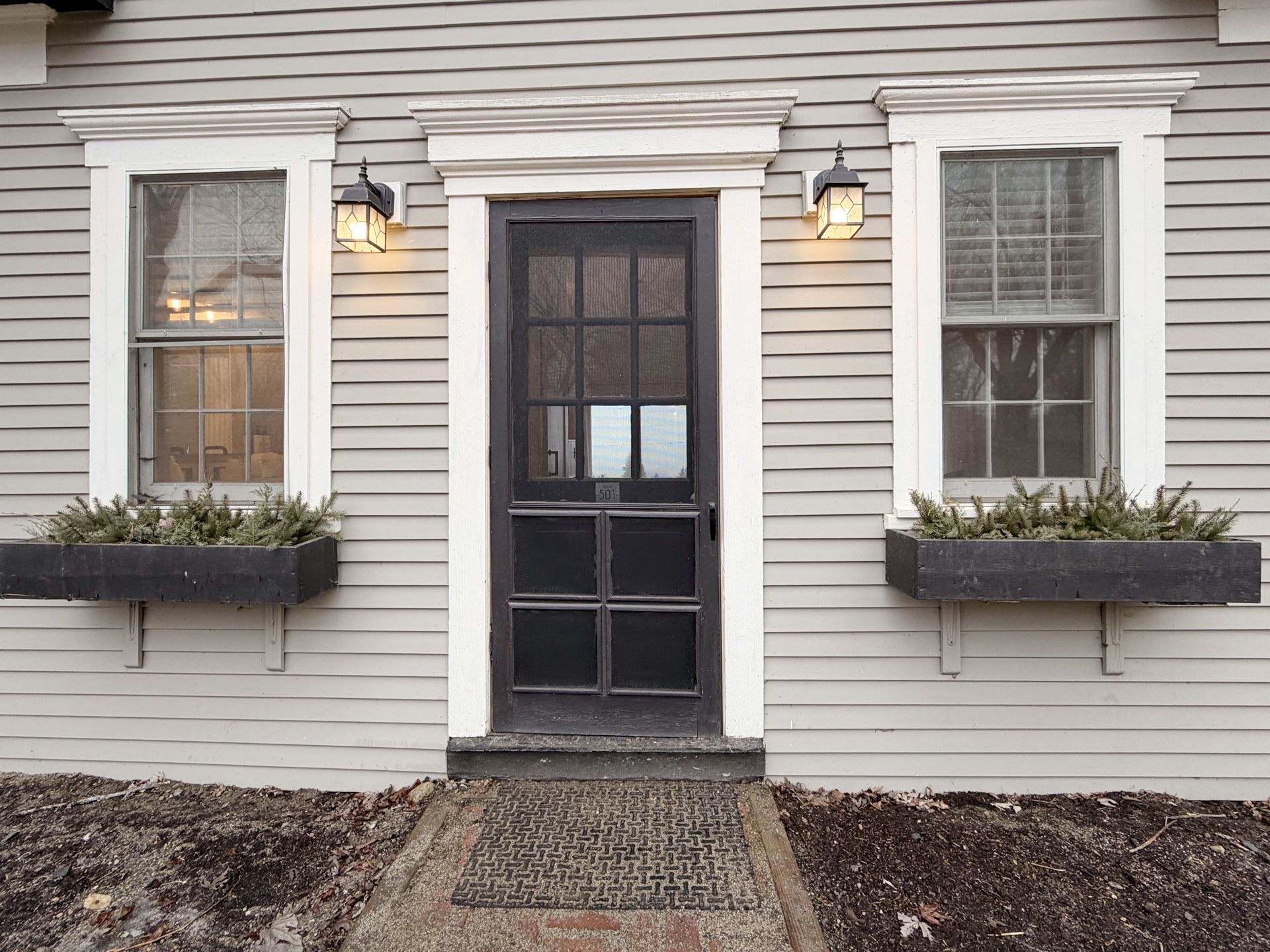 Front entrance of a house with a black door, white trim, and two flower boxes under lit wall lamps