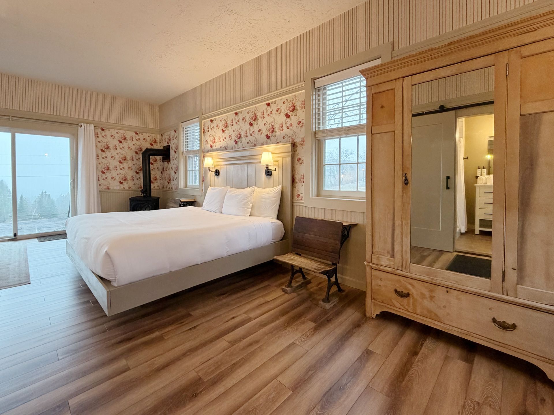 Bright bedroom with a white bed, floral wallpaper, wood floors, and a large wooden wardrobe.