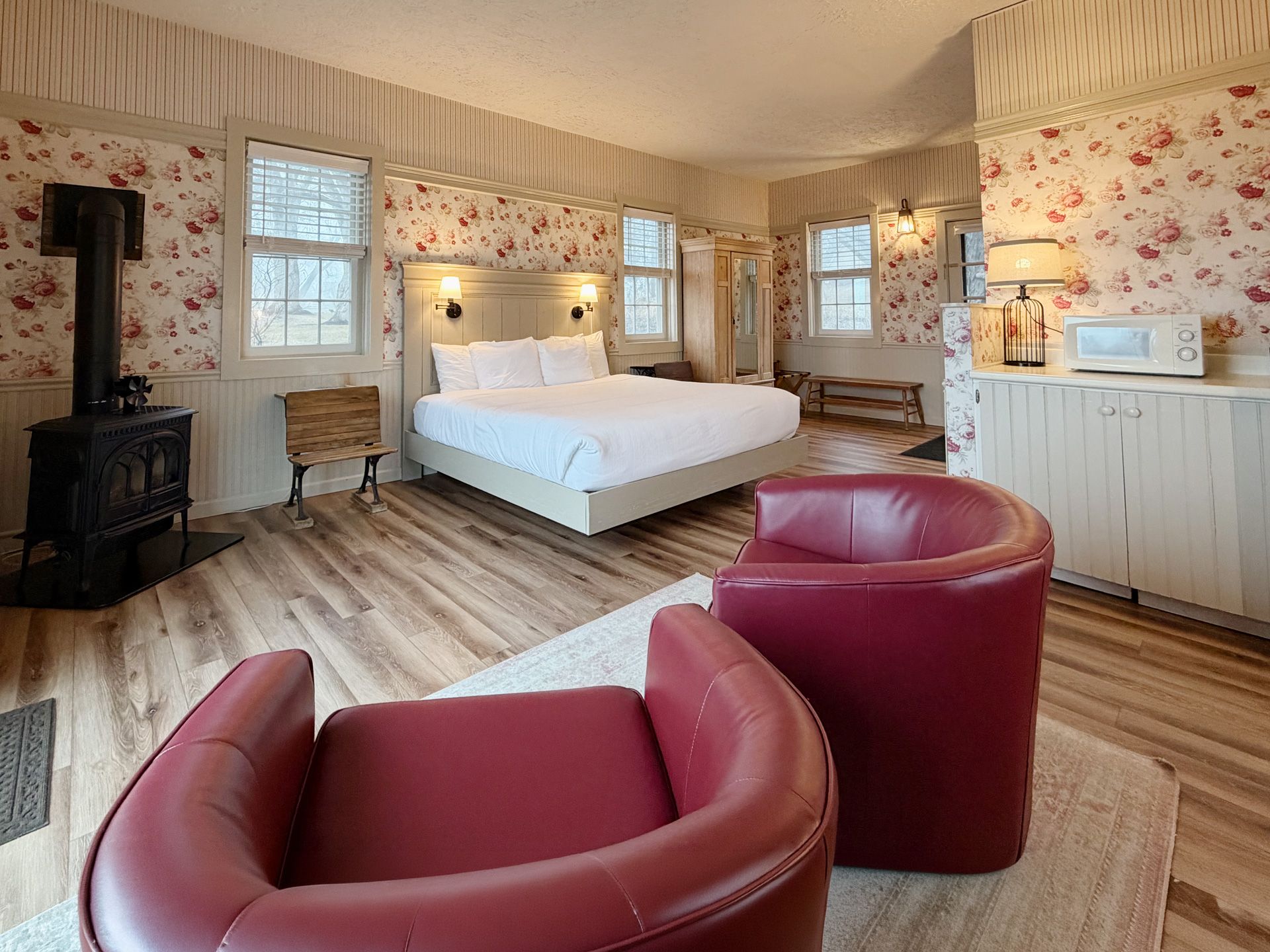 Cozy bedroom with floral wallpaper, white bed, red chairs, wood floors, and a black stove heater