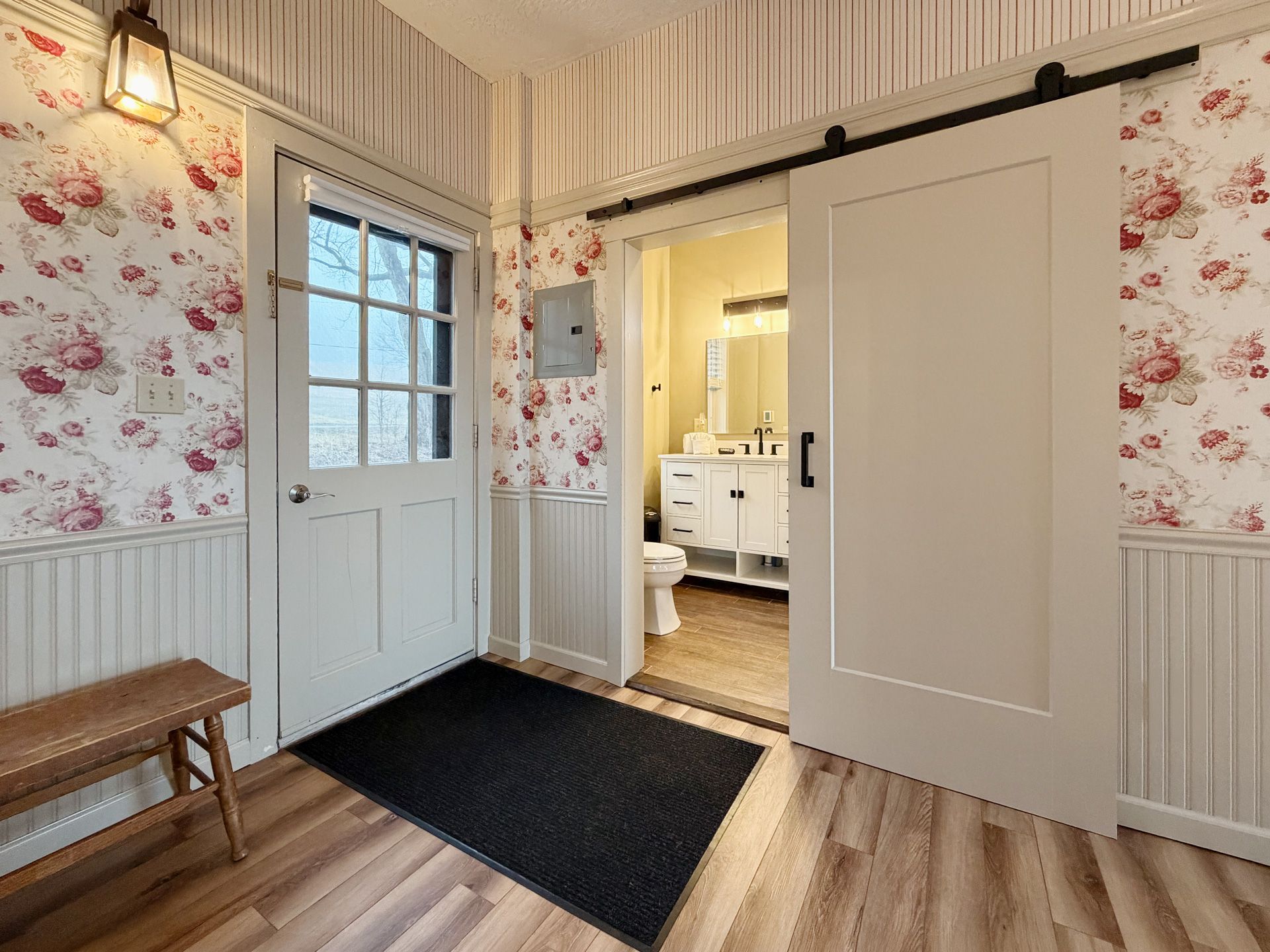 Cozy entryway with floral wallpaper, white doors, wood floor, black rug, and bench under a hanging light.