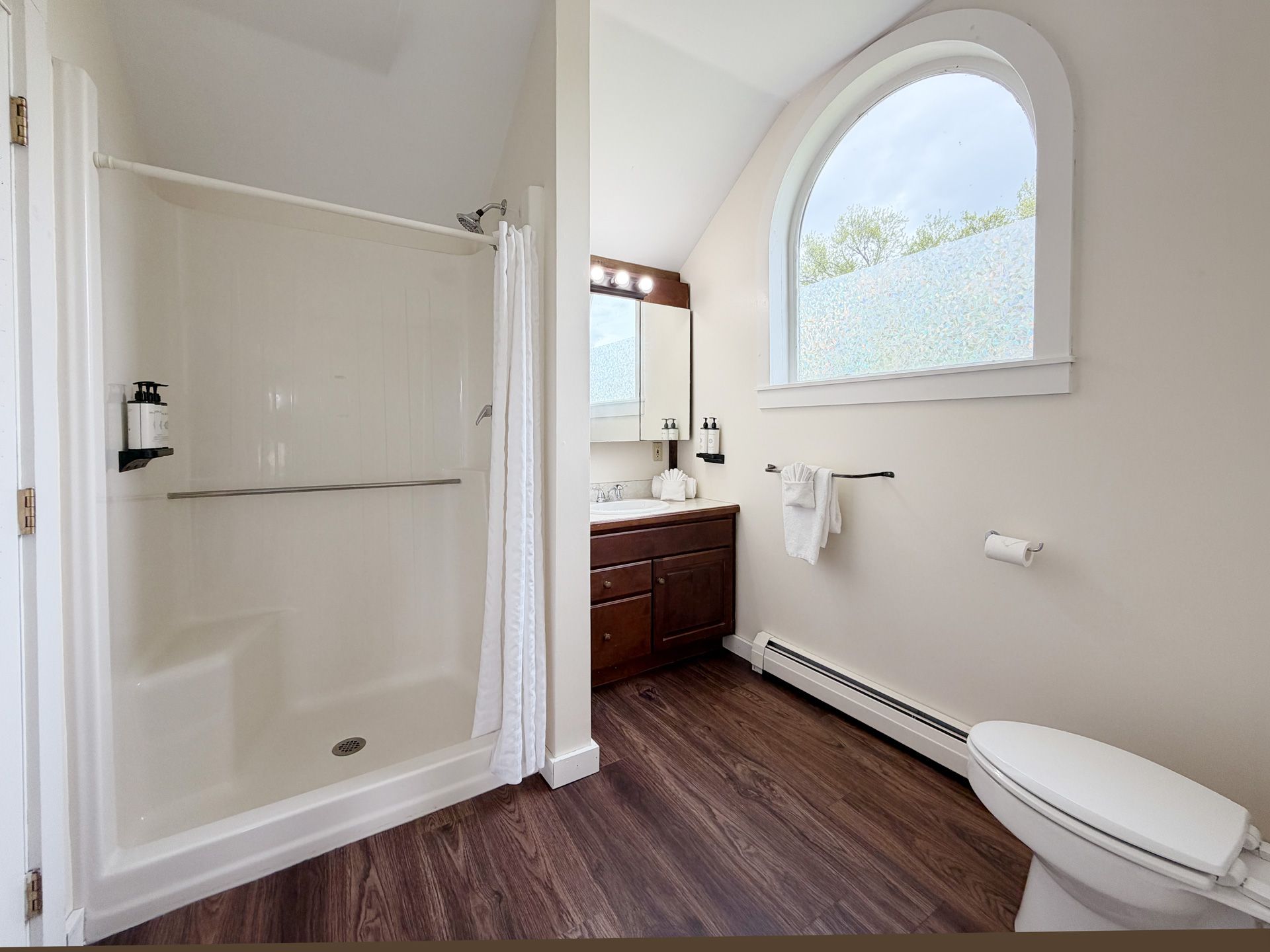 Modern bathroom with a shower, vanity, arched window, and toilet on wood-look flooring