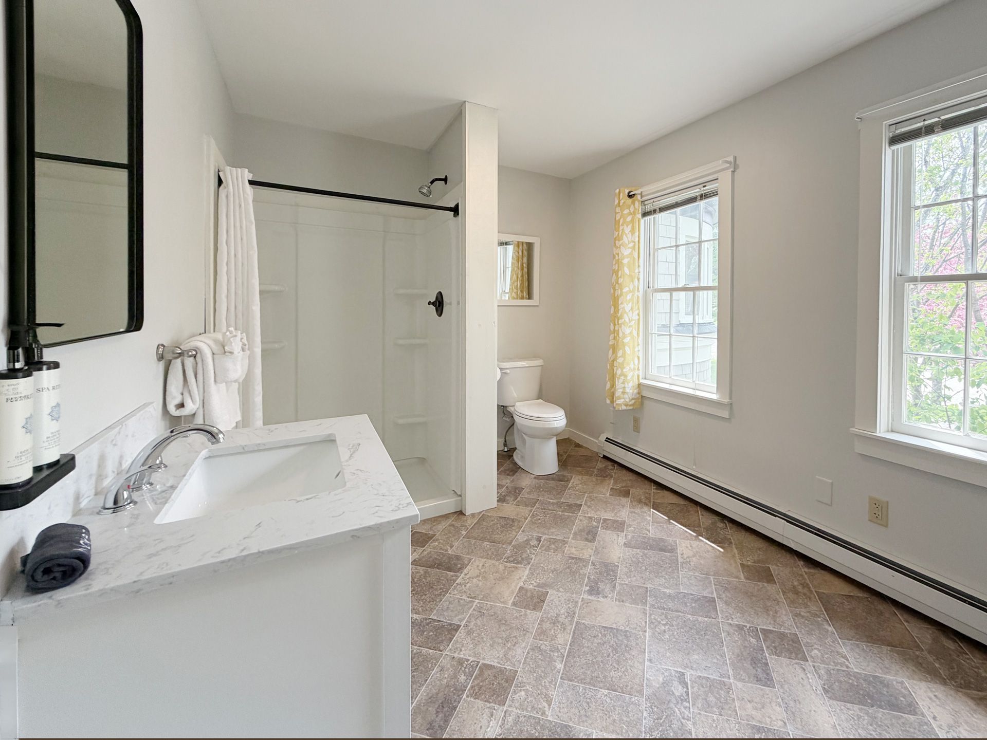 Bright bathroom with double vanity, walk-in shower, toilet, and two windows on a tiled floor.