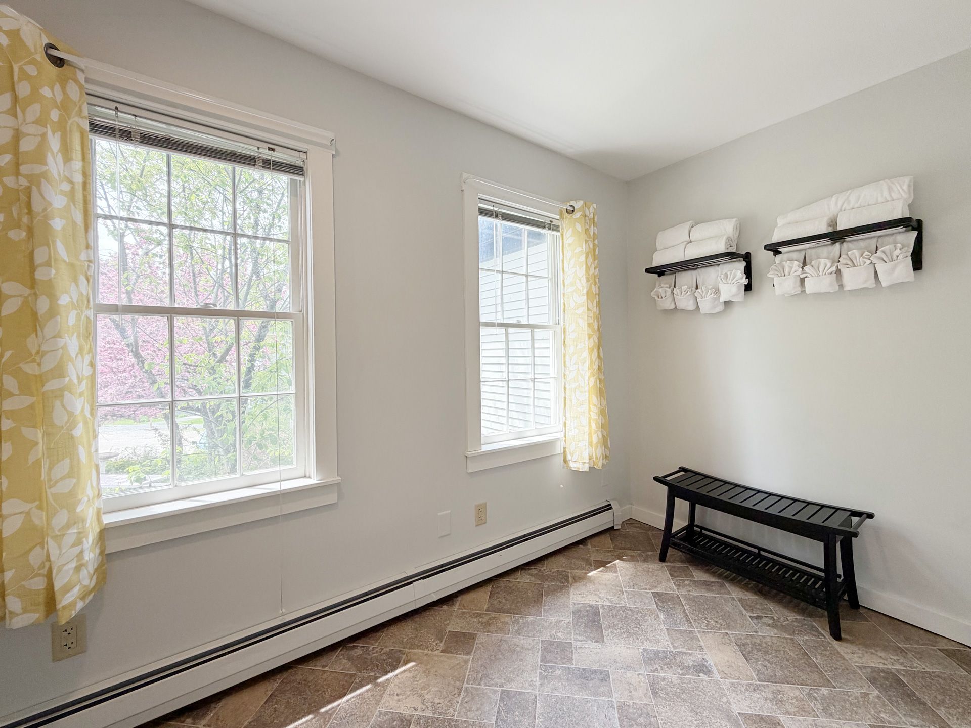 Empty bright room with two windows, white walls, beige carpet, and a black bench on the right