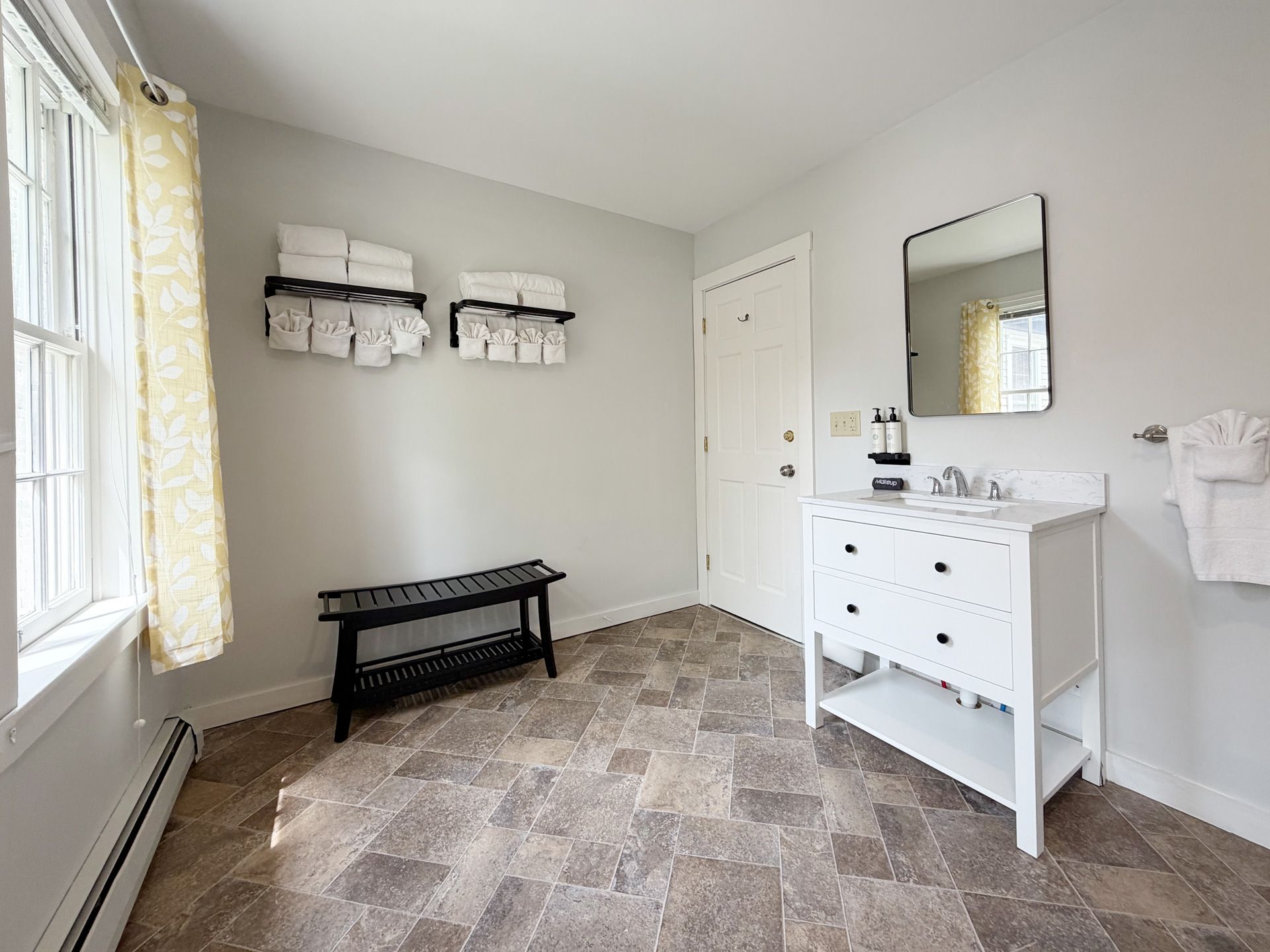 Bright neutral room with a bench, small white vanity, wall shelves, mirror, and window curtains