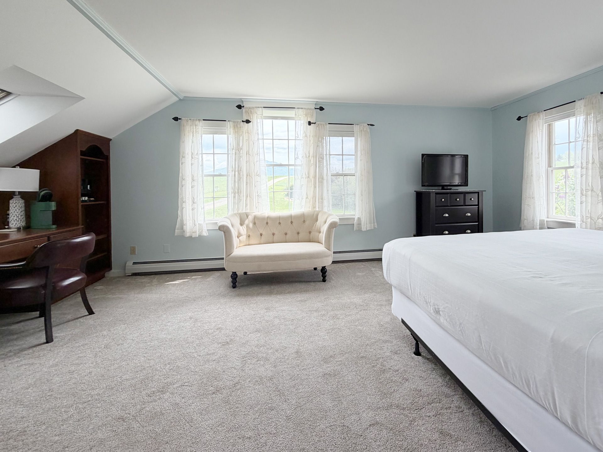Bright bedroom with large windows, gray carpet, white bed, chaise lounge, desk, and TV stand