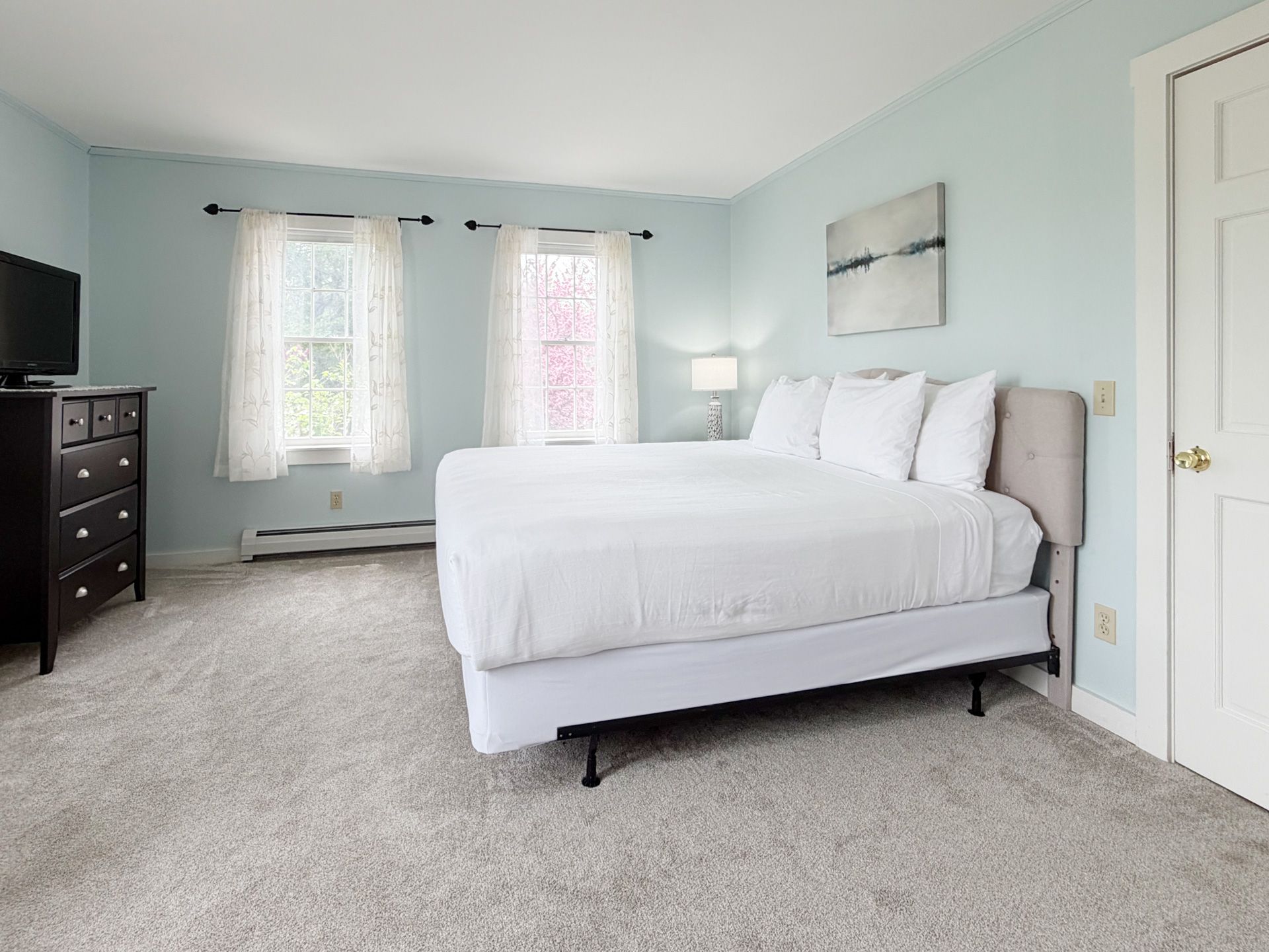 Bright bedroom with a large white bed, light blue walls, gray carpet, and windows with curtains