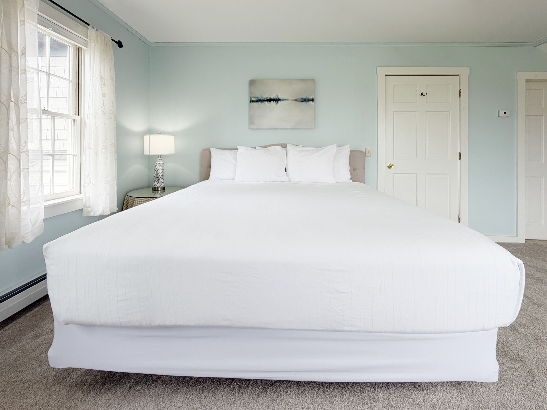 Bright minimalist bedroom with a large white bed, pale blue walls, window, lamp, and closed doors