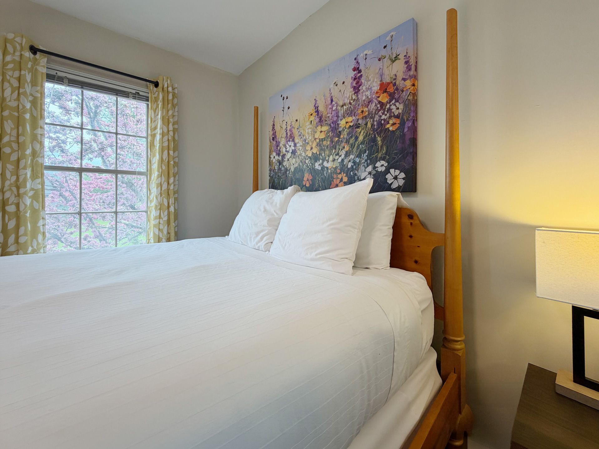 Bright bedroom with a neatly made white bed, floral wall art, and a window with patterned curtains