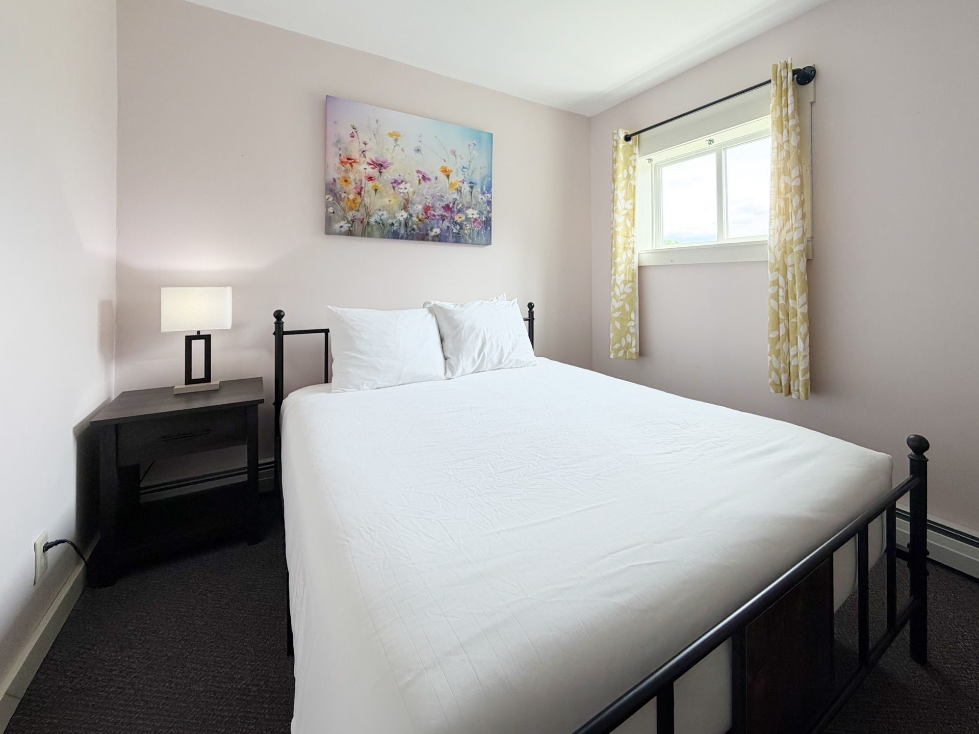 Small bedroom with a black bed frame, white bedding, nightstand lamp, and window with curtains.