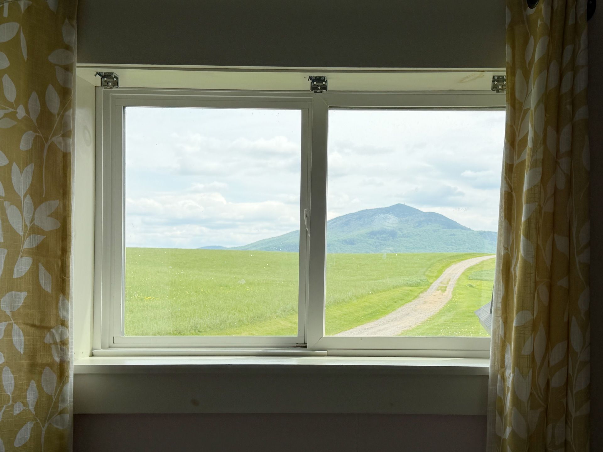 Window view of green fields, a dirt road, and a distant mountain under a cloudy sky