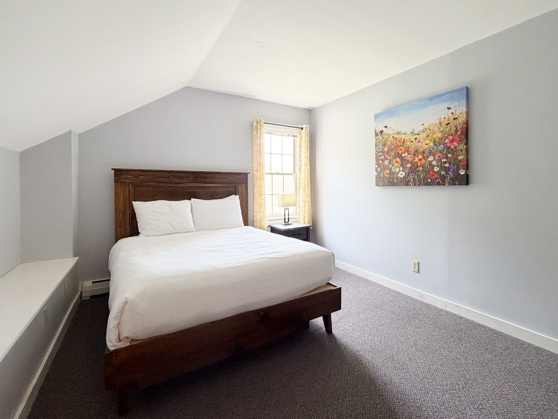 Bright bedroom with a wooden bed, gray carpet, white walls, and a colorful painting.