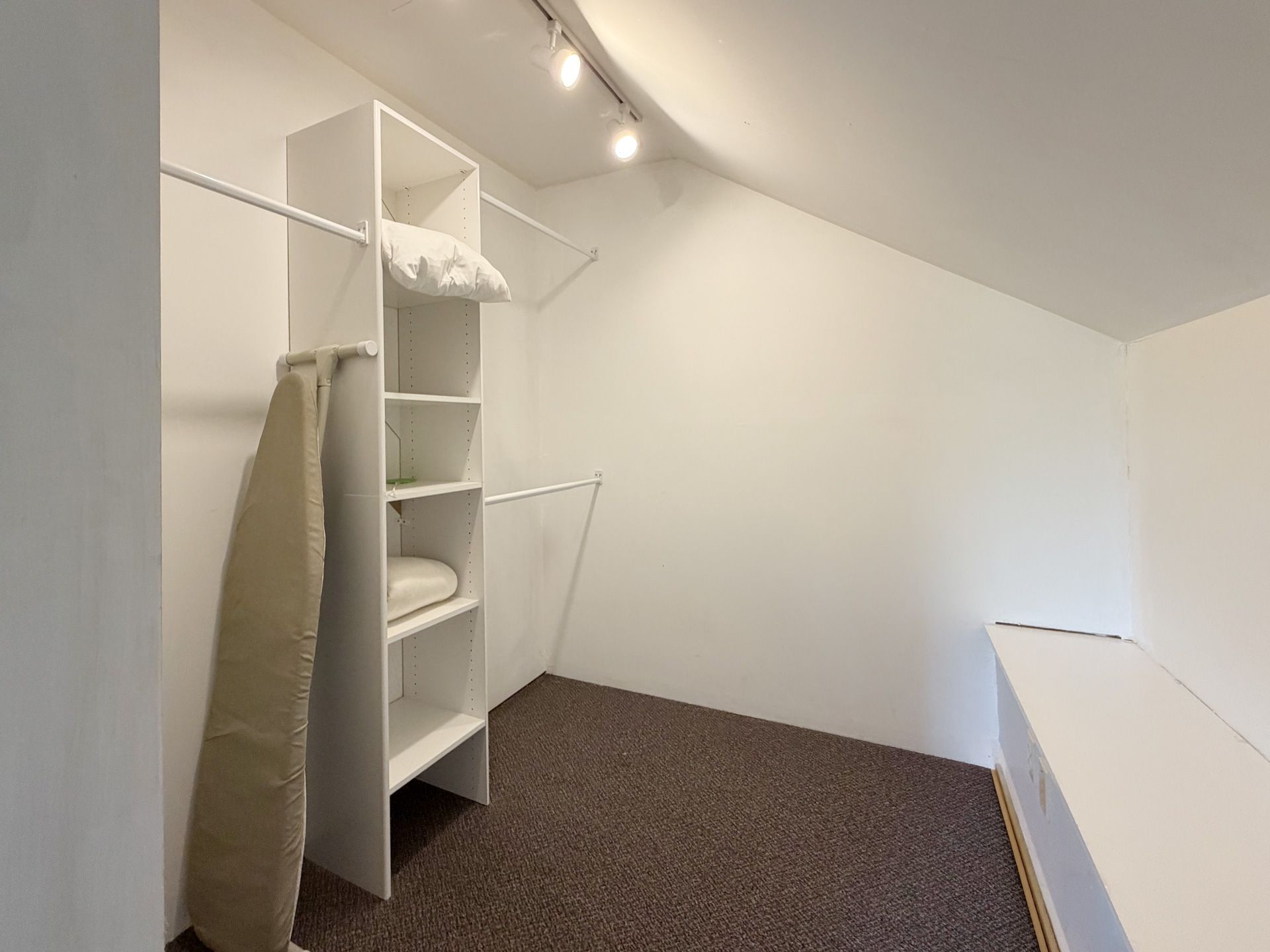 Small walk-in closet with white shelves, hanging rod, and brown carpet under a sloped ceiling.