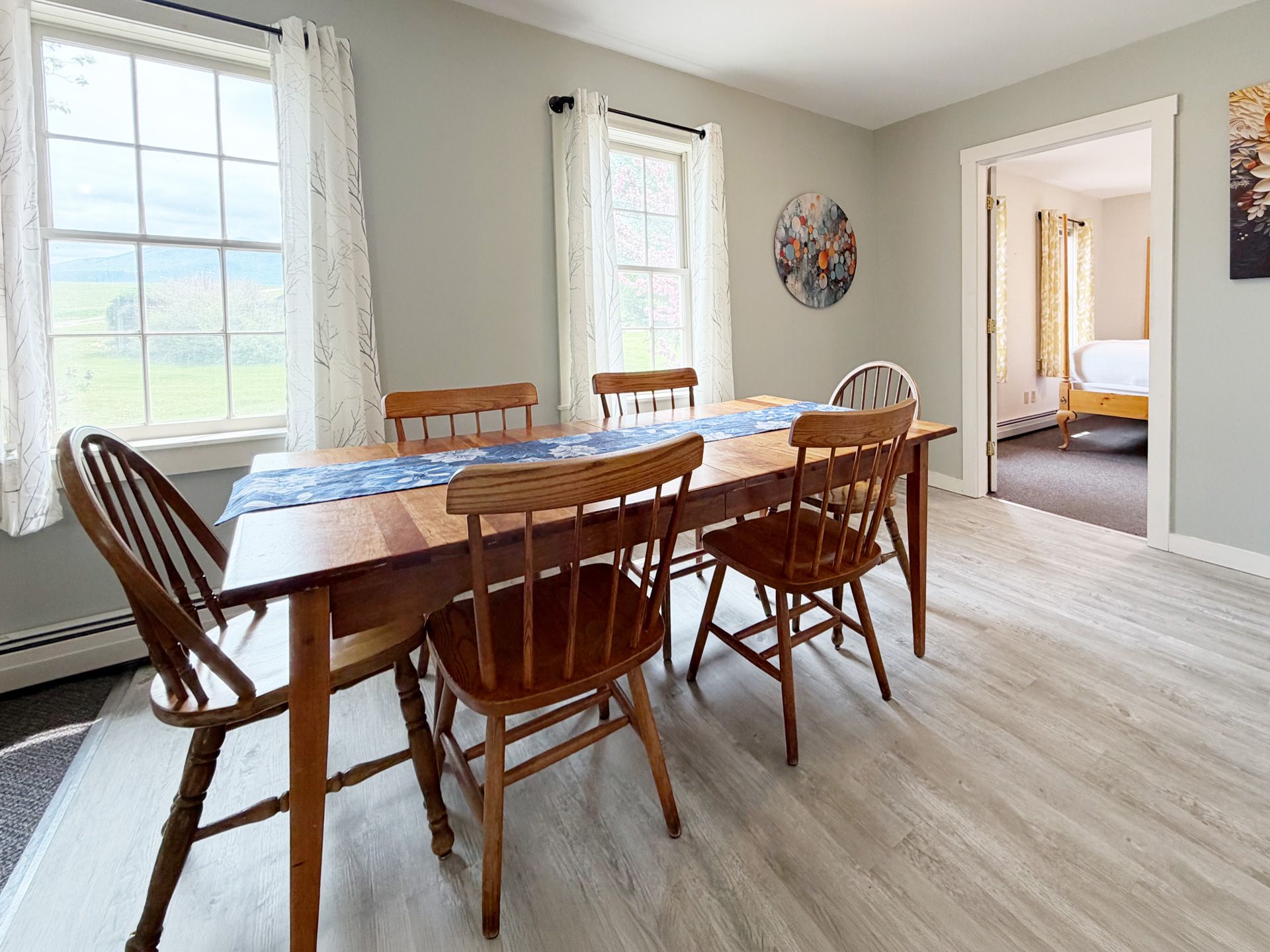 Bright dining room with wooden table and chairs, white curtains, and an open doorway to a bedroom.