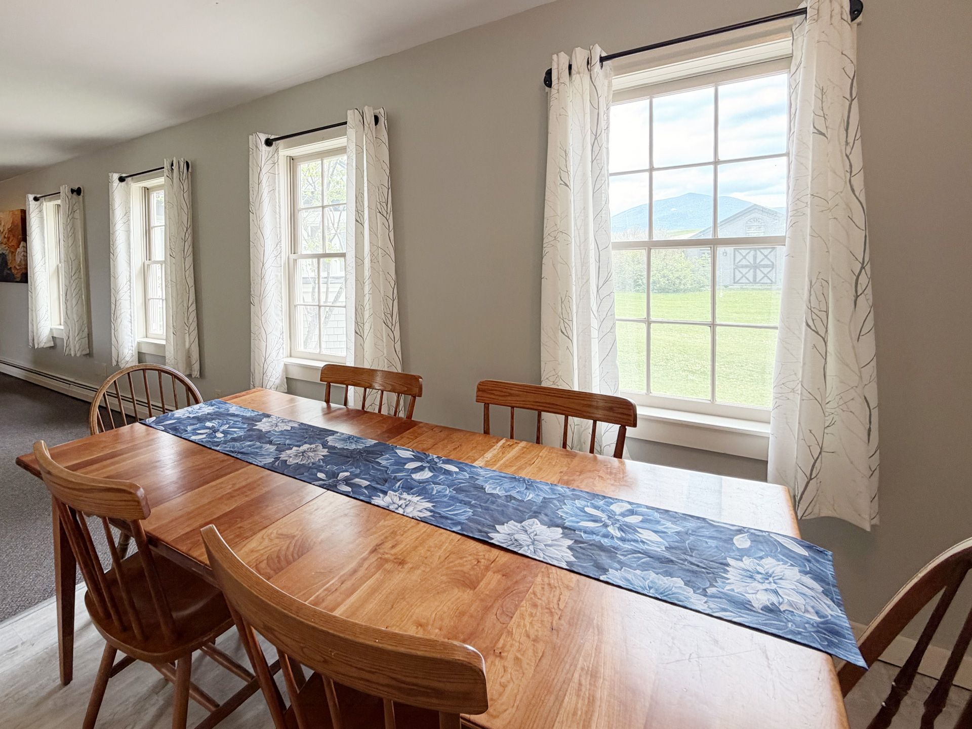 Dining room with wooden table and chairs, blue runner, and large windows with lace curtains