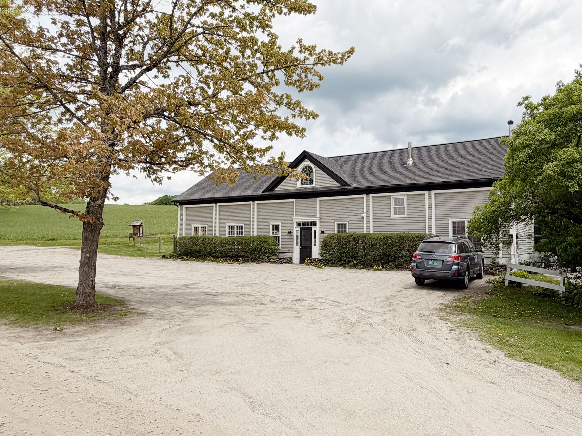 Gravel driveway leading to a gray house with parked cars under cloudy skies.