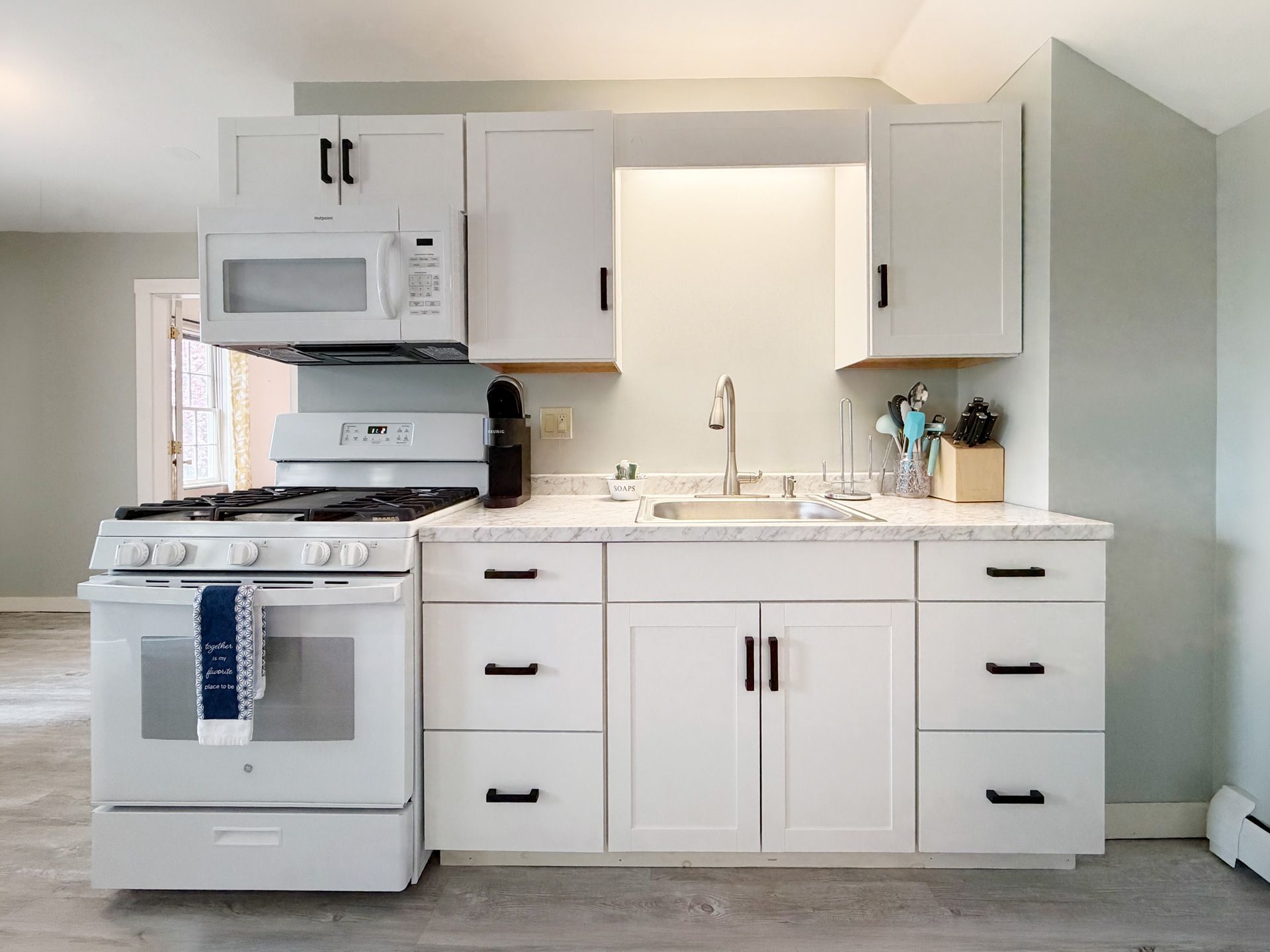 Bright white compact kitchen with stove, microwave, sink, cabinets, and black hardware.