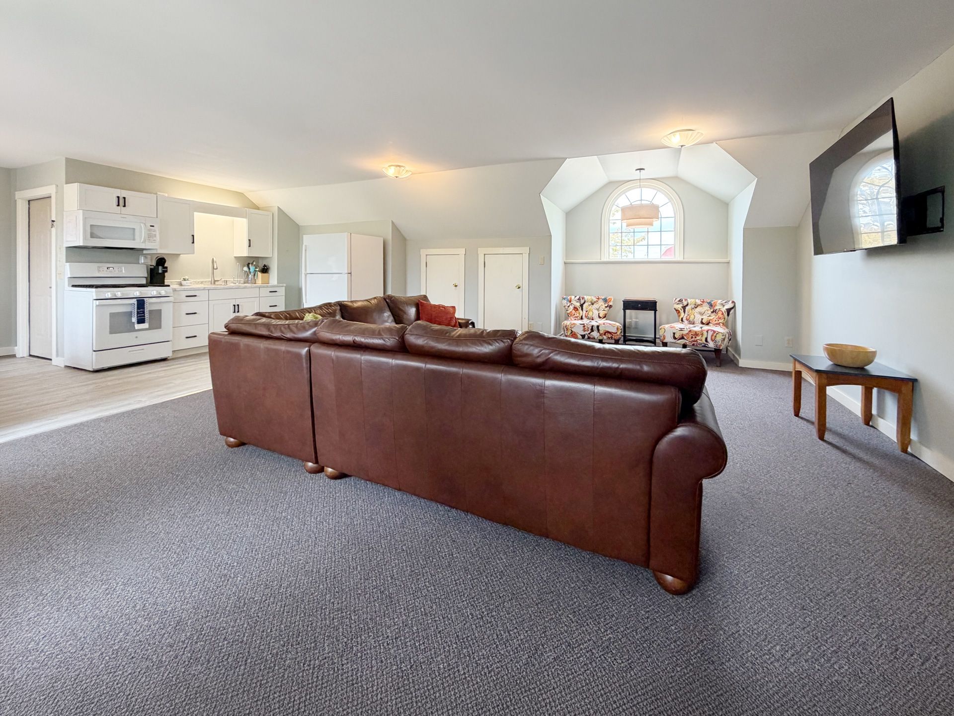 Bright open living room with brown leather sofa, white kitchen, wall TV, and fireplace alcove