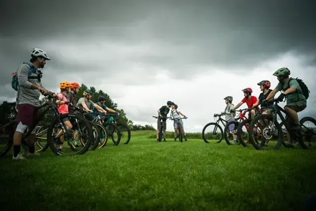 Cyclists lined up on a grassy field under dark, cloudy skies, preparing to start a race