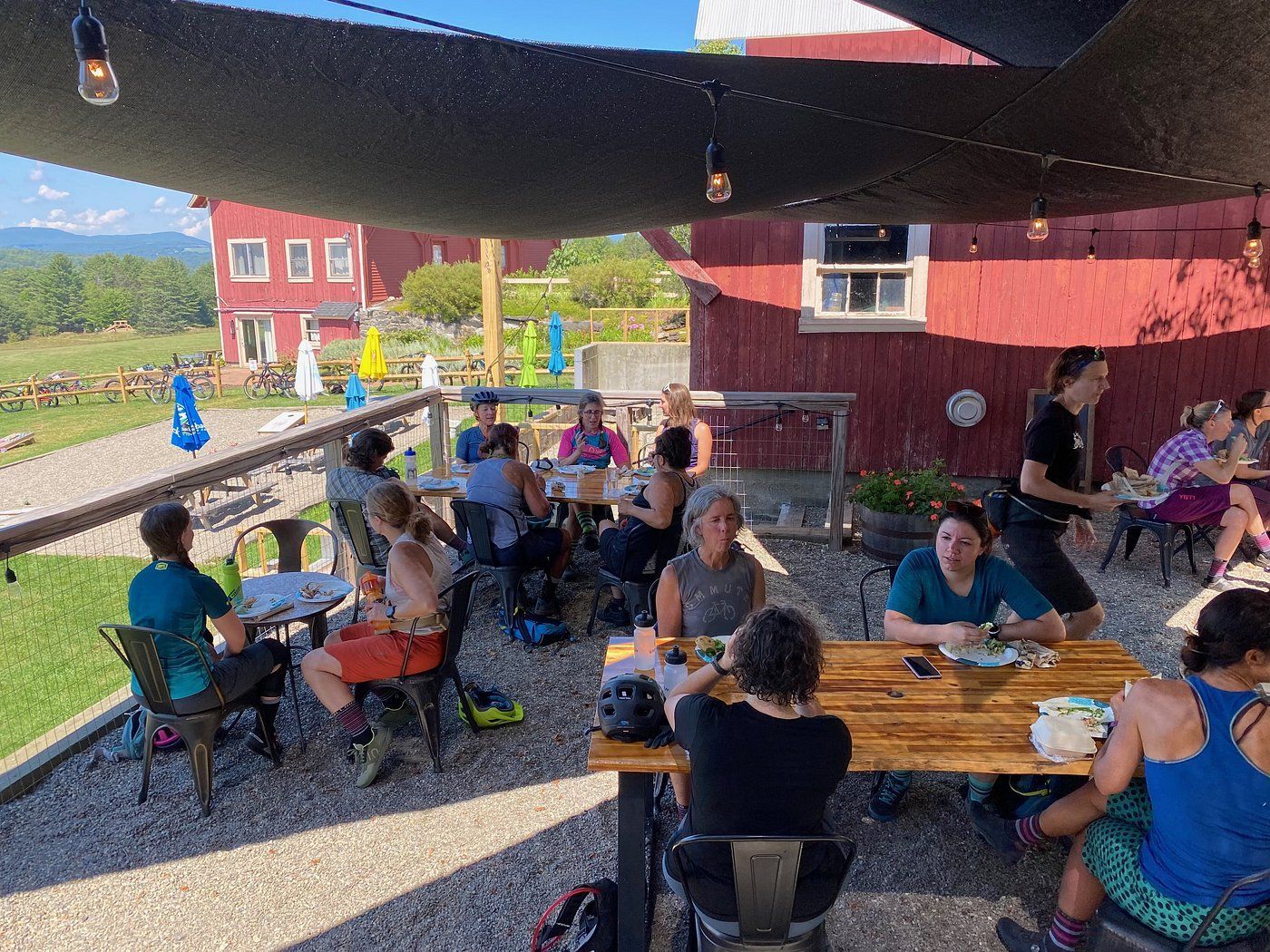 People seated at picnic tables under a shade canopy outside a red building on a sunny day