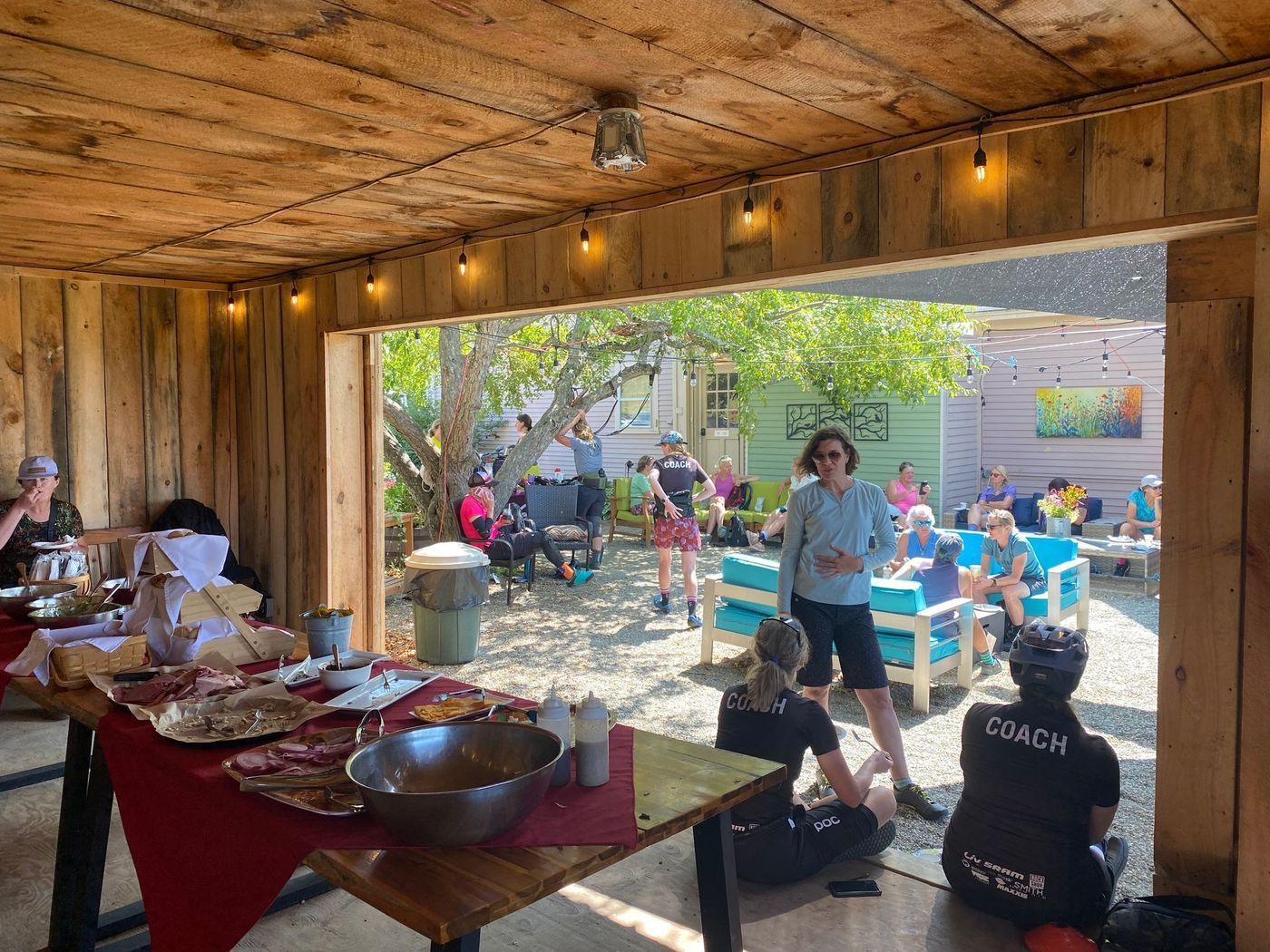 Outdoor backyard party under a wooden patio, with a buffet table and guests seated at tables in the yard.