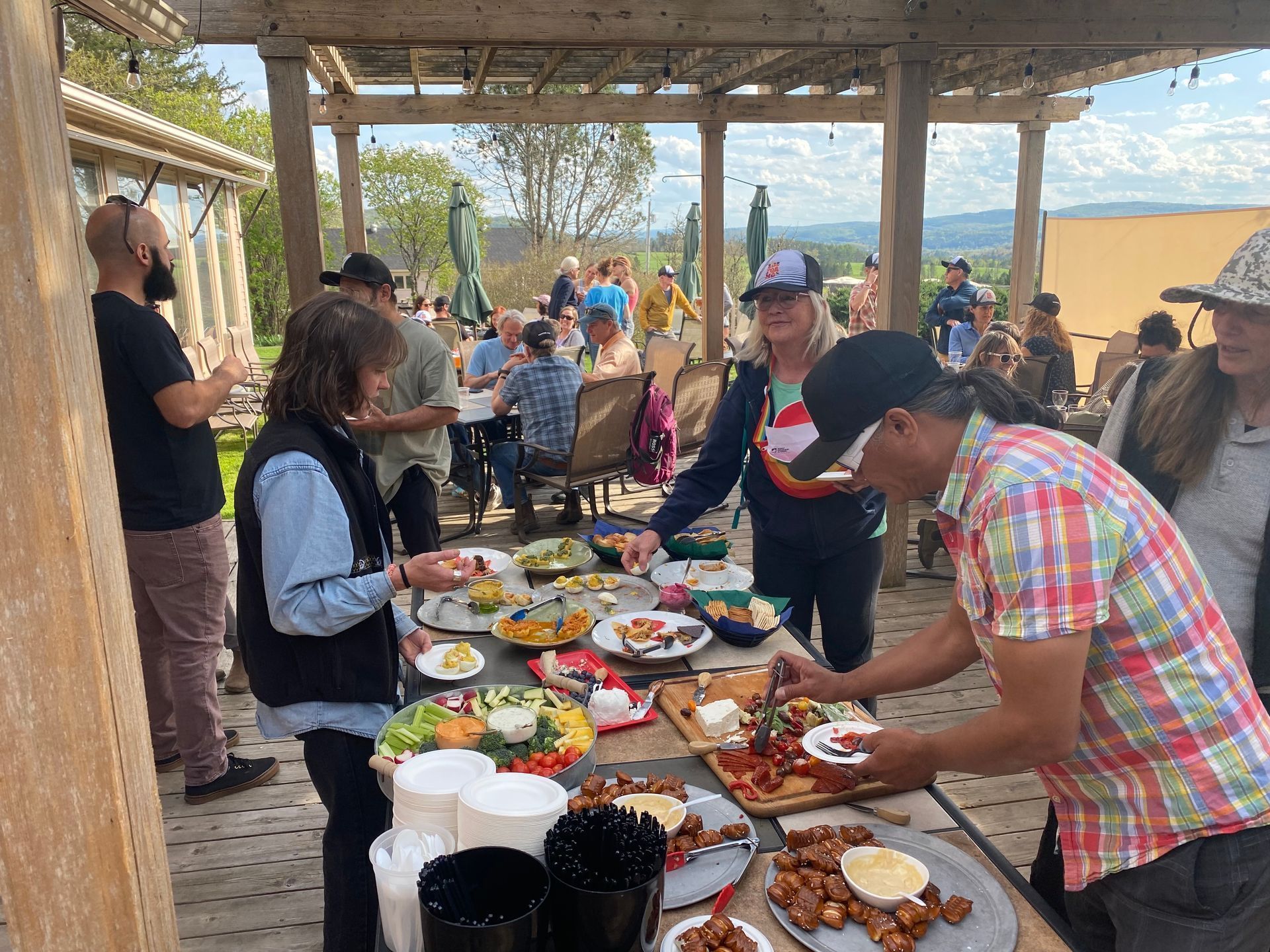 People gathered under a pavilion around a long table of food, with a beach visible in the background.