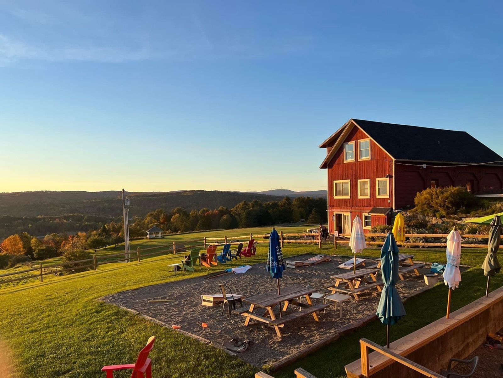 Sunlit patio with tables and umbrellas beside a red house overlooking a scenic valley.