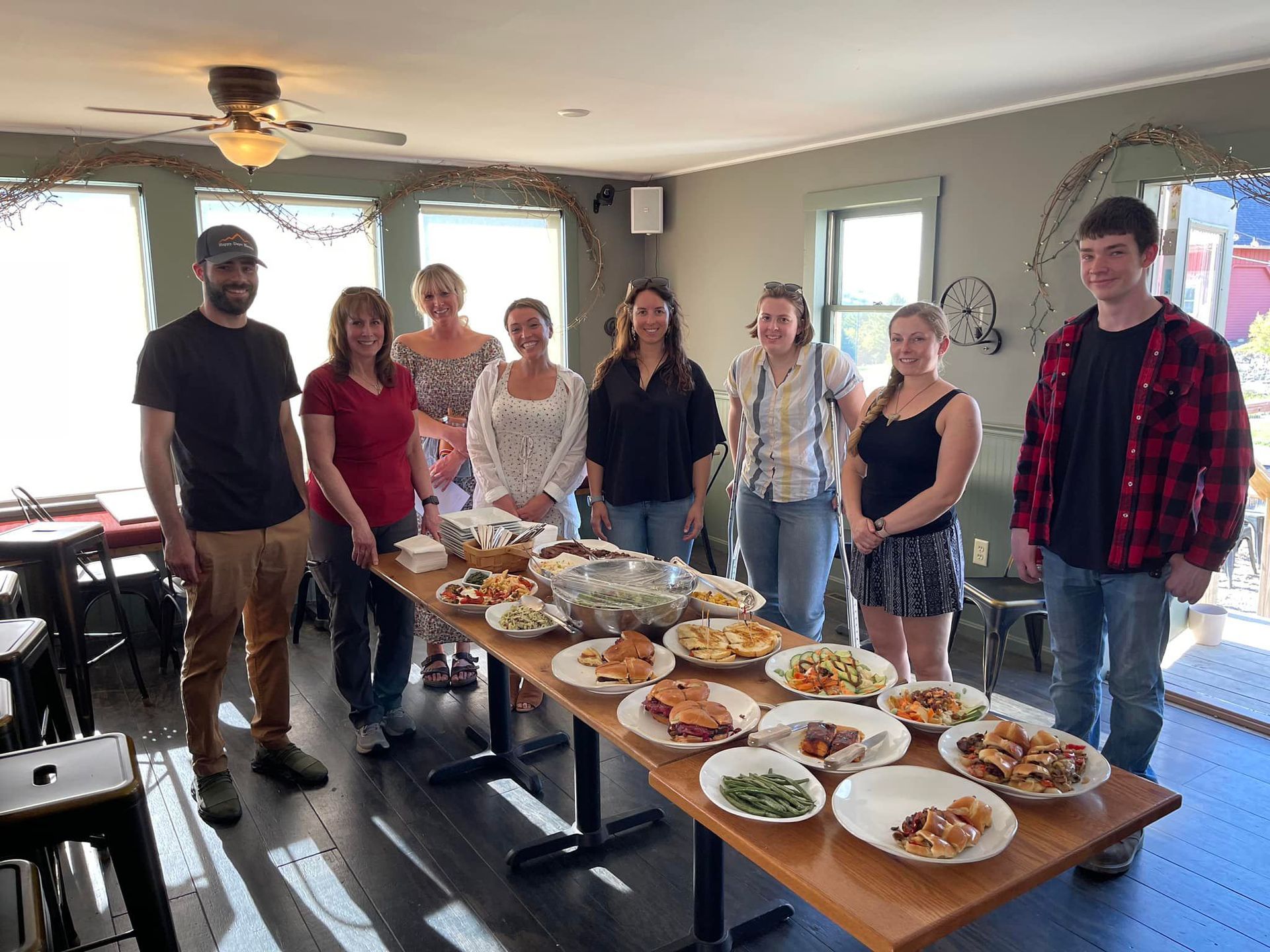 Group of people standing behind a long table of dishes in a bright living room, smiling at the camera
