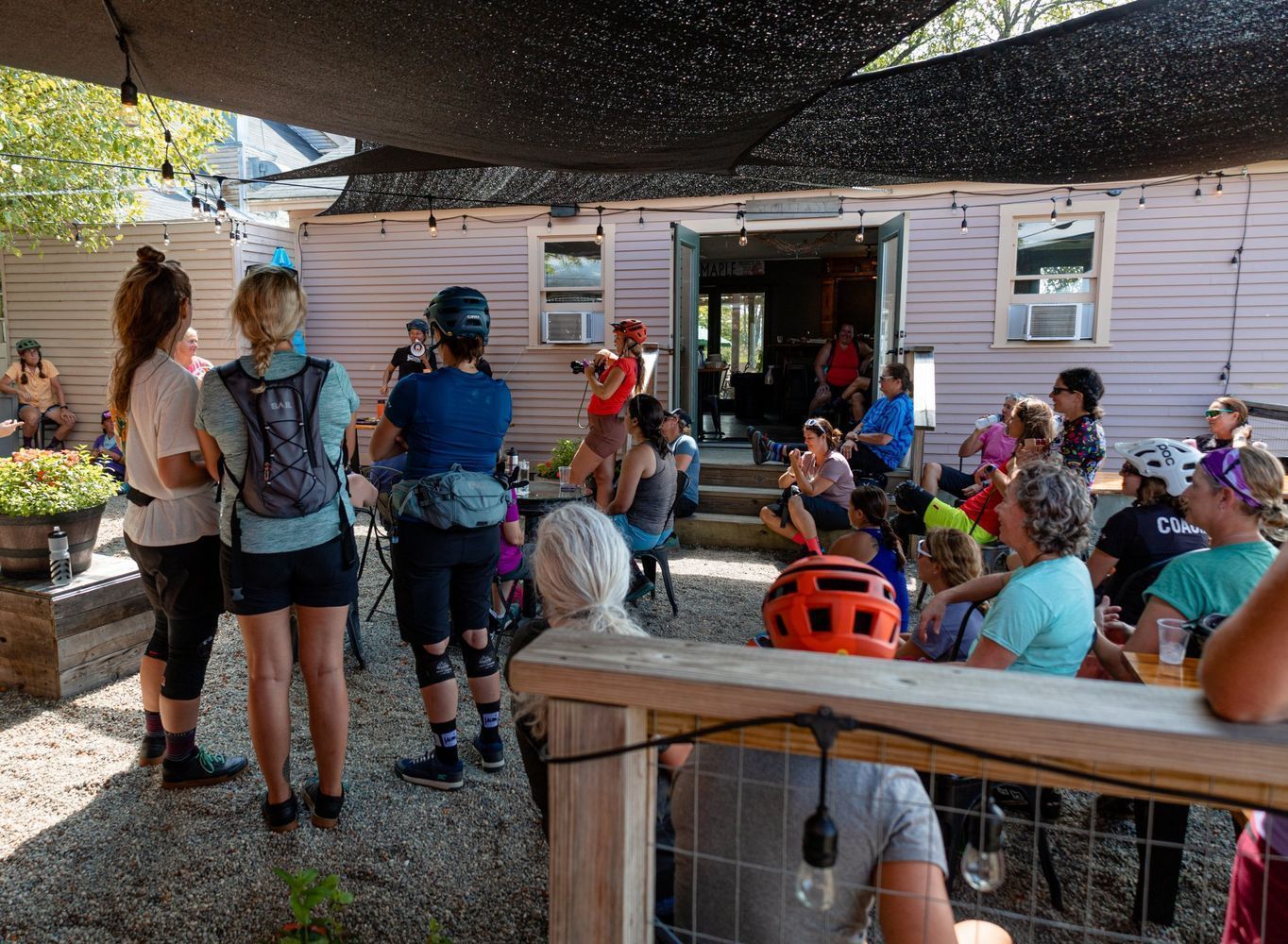 People seated and standing at an outdoor gathering under a shade canopy beside a white building.