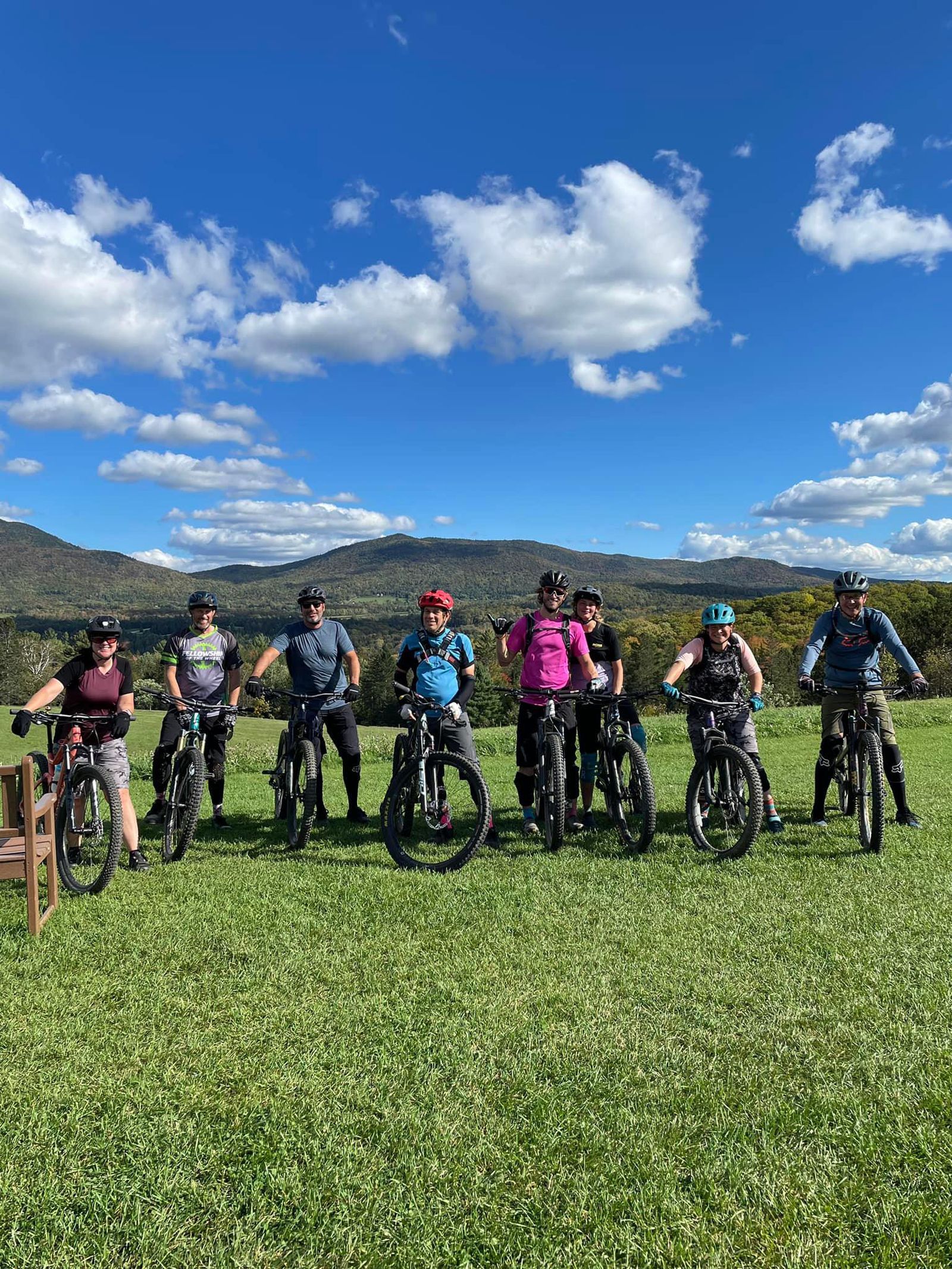 Cyclists posing with mountain bikes in a grassy field under a blue sky with clouds, mountains in the background