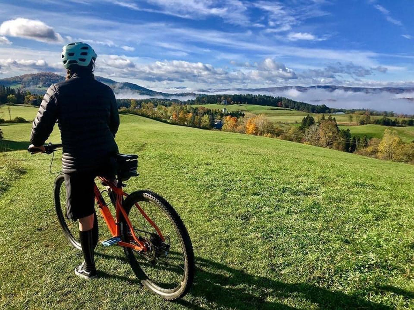 Cyclist in a teal helmet standing on a red bike overlooking a sunny green hillside and valley