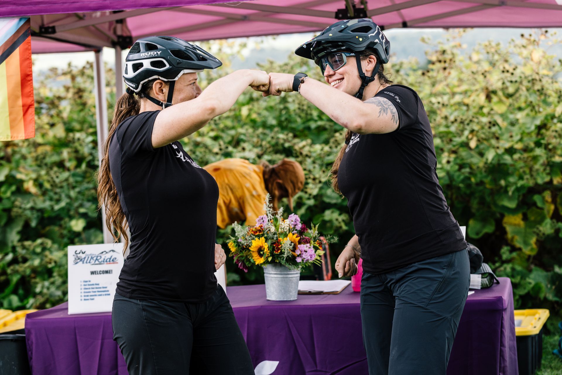 Two cyclists fist-bump at an outdoor event booth with a purple tablecloth and flowers.