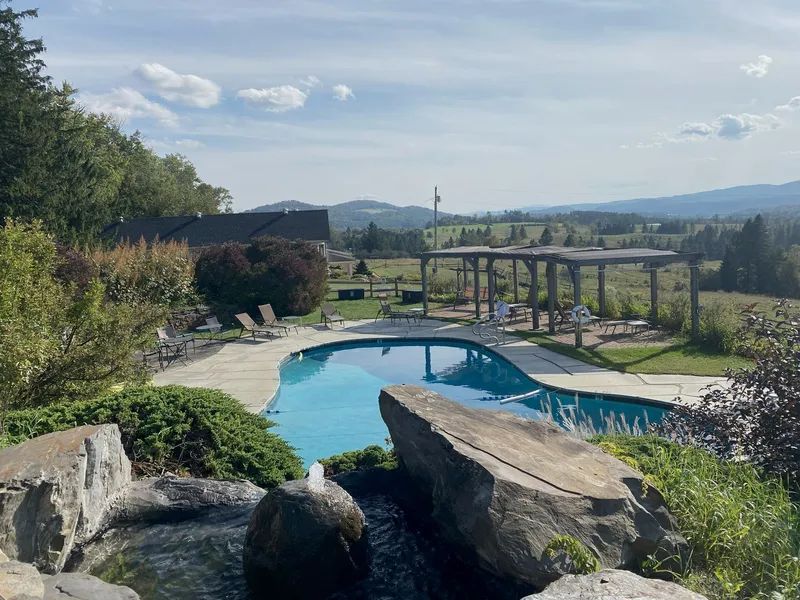 Outdoor pool with blue water, stone rocks in foreground, and a pergola overlooking a scenic hillside view