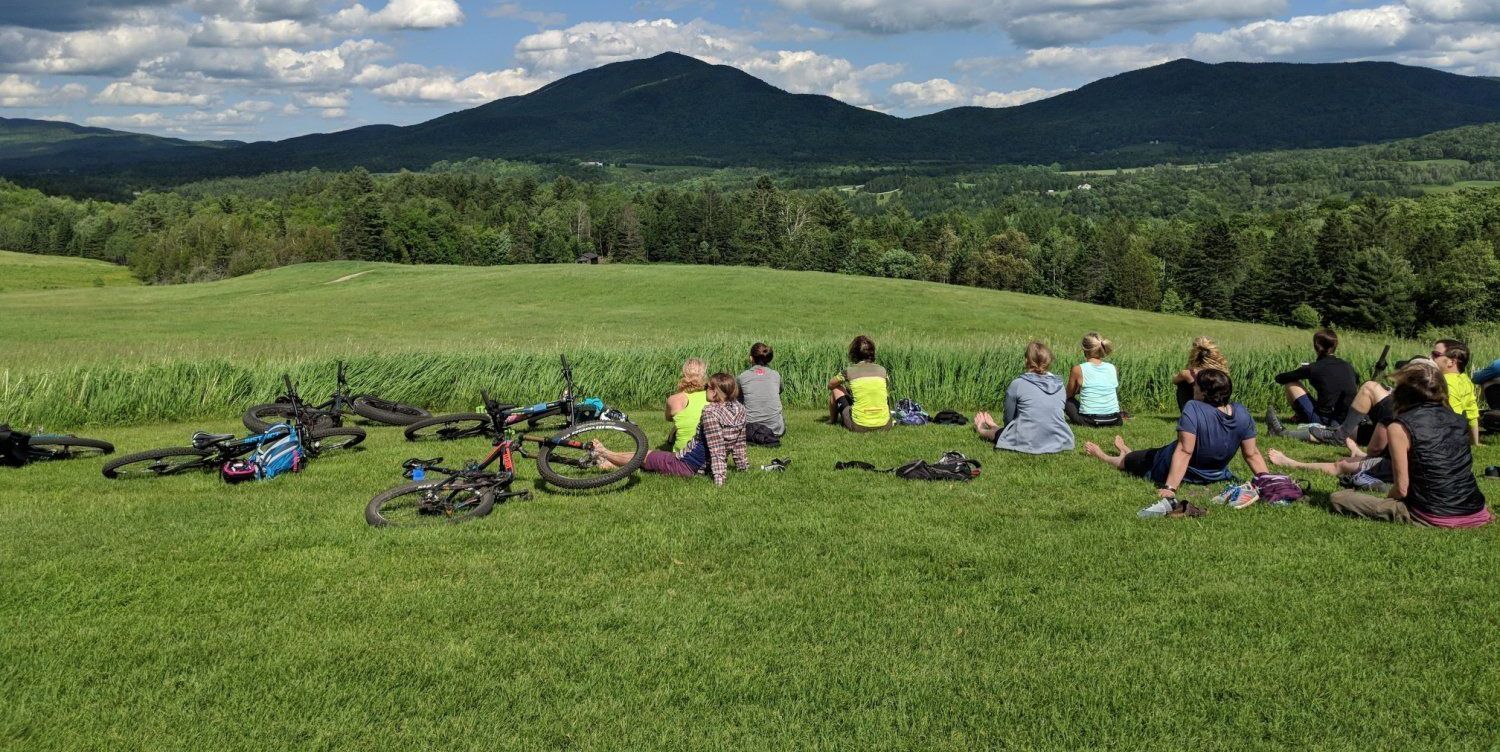 Cyclists resting on a grassy hill with bikes, overlooking forested mountains and blue sky.