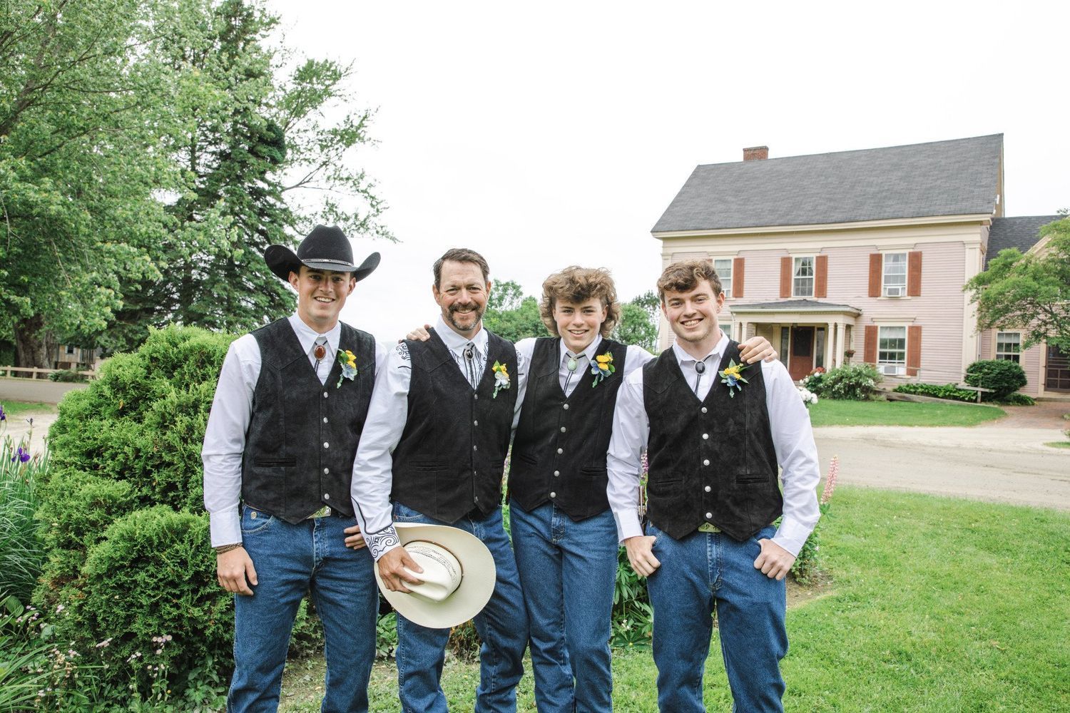 Four men in matching vests and jeans stand outdoors in front of a house, one holding a hat.