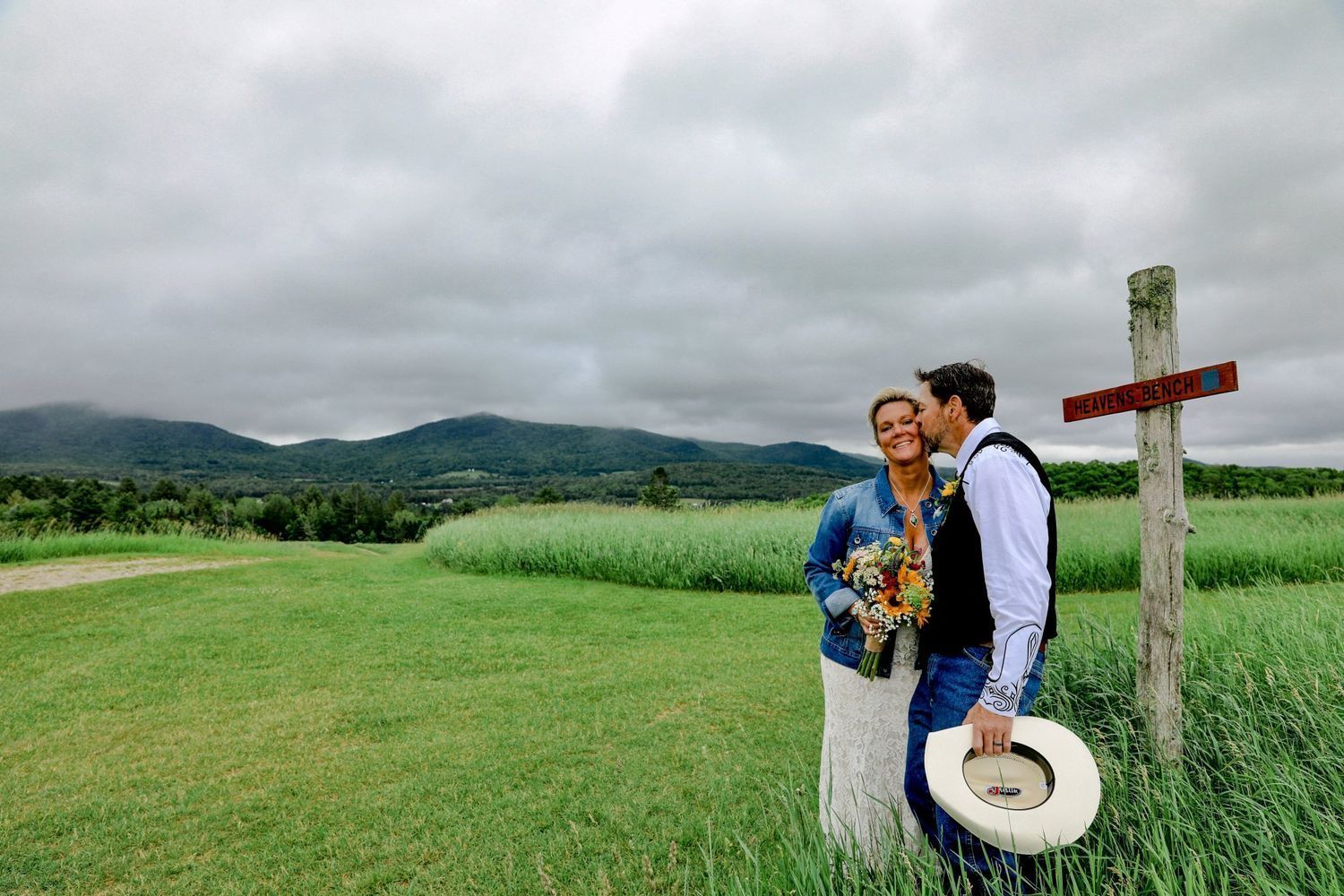 Couple standing by a wooden cross in a green field, with mountains and cloudy sky behind them.