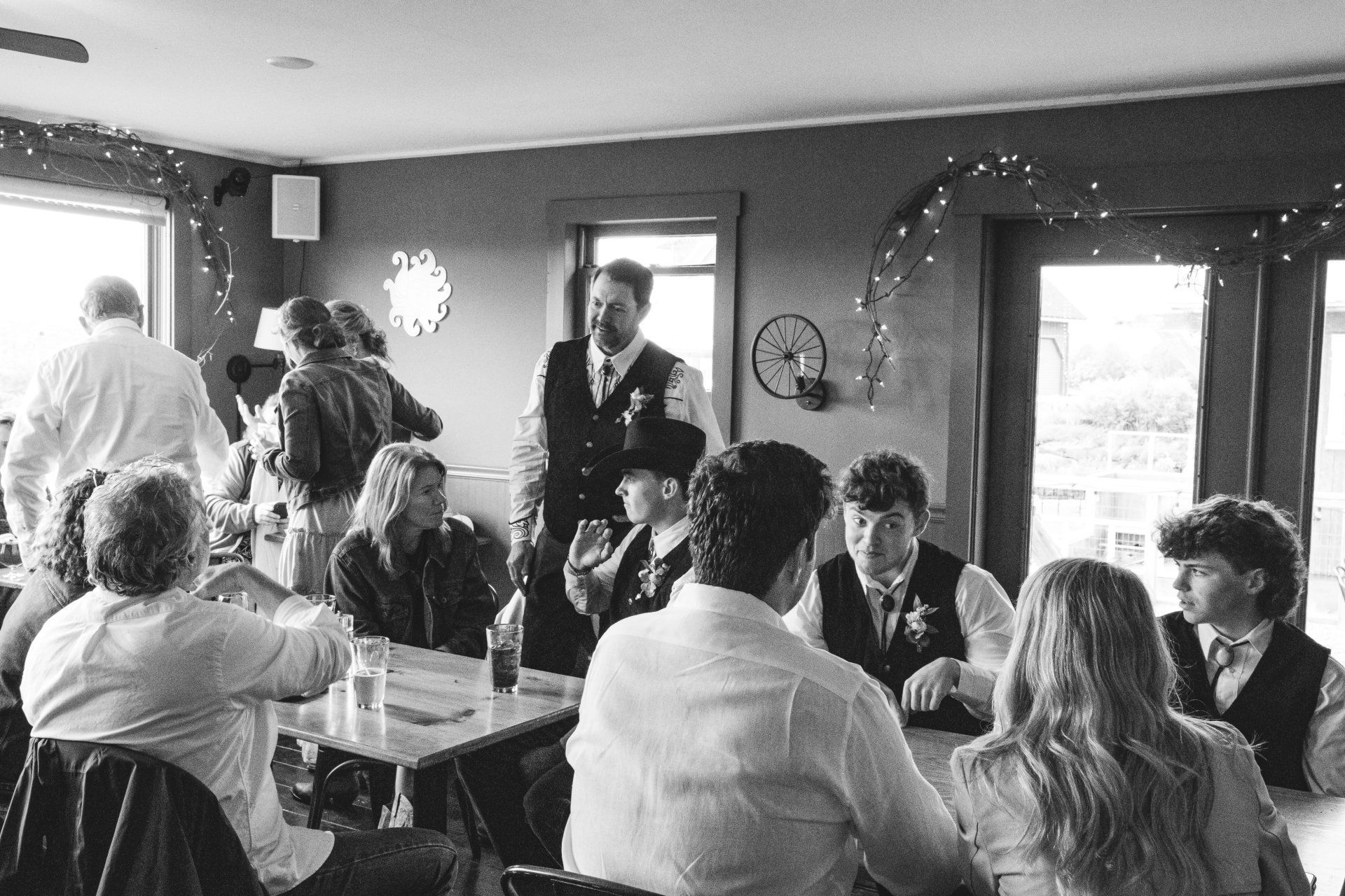 Black-and-white wedding reception with guests seated around tables as a man gives a speech.