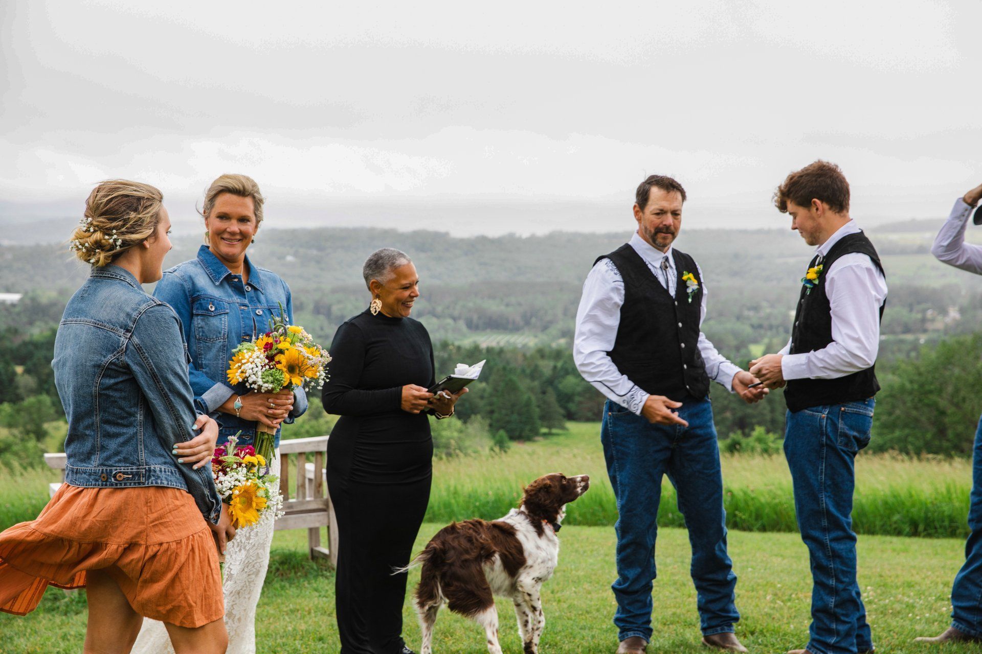 Five people and a dog standing in a grassy field, chatting near a scenic hillside.