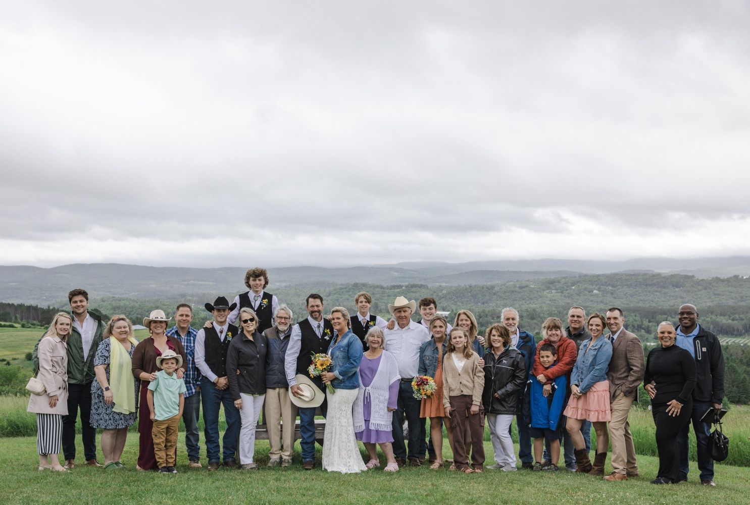 Large group posing outdoors on a grassy hill under a cloudy sky, with mountains in the background.